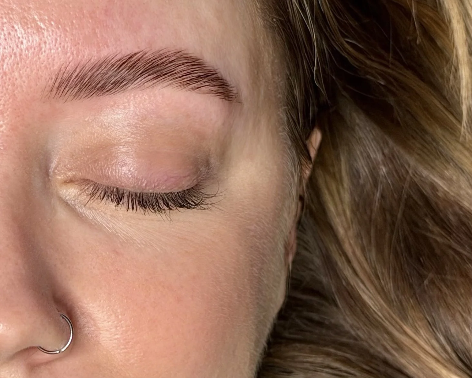 Close-up of a person's face with a focus on their eyebrow, eyelash, and closed eye, alongside their nose and a glimpse of a nose ring, with a background of brown hair.