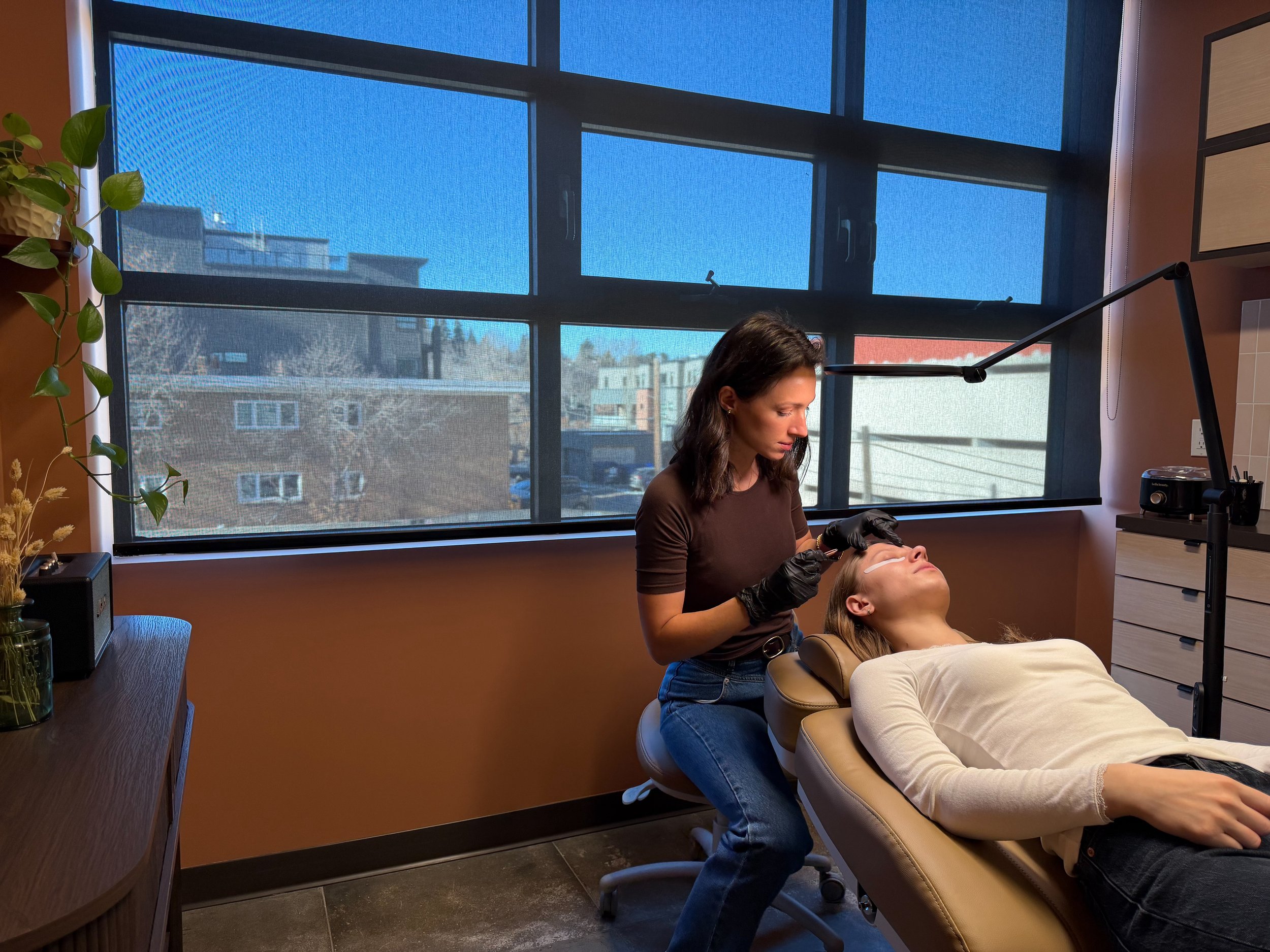 A woman with dark hair in a brown shirt and black gloves is performing a cosmetic treatment on a woman lying on a beige chair in a spa or clinic room near a large window with a view of buildings and trees outdoors.