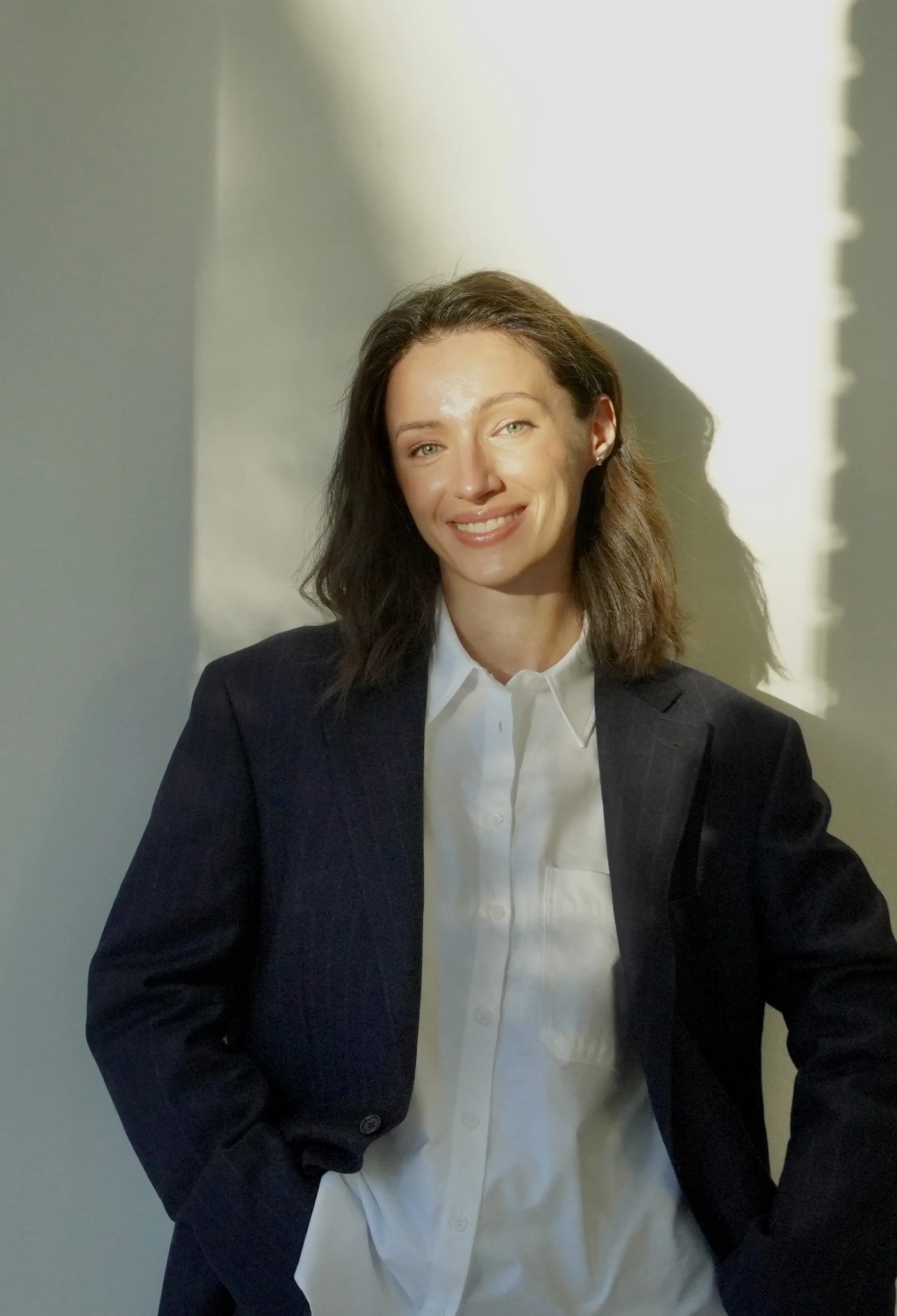 A confident woman with shoulder-length brown hair wearing a black blazer and white shirt, smiling, standing against a light-colored wall with sunlight casting a shadow.