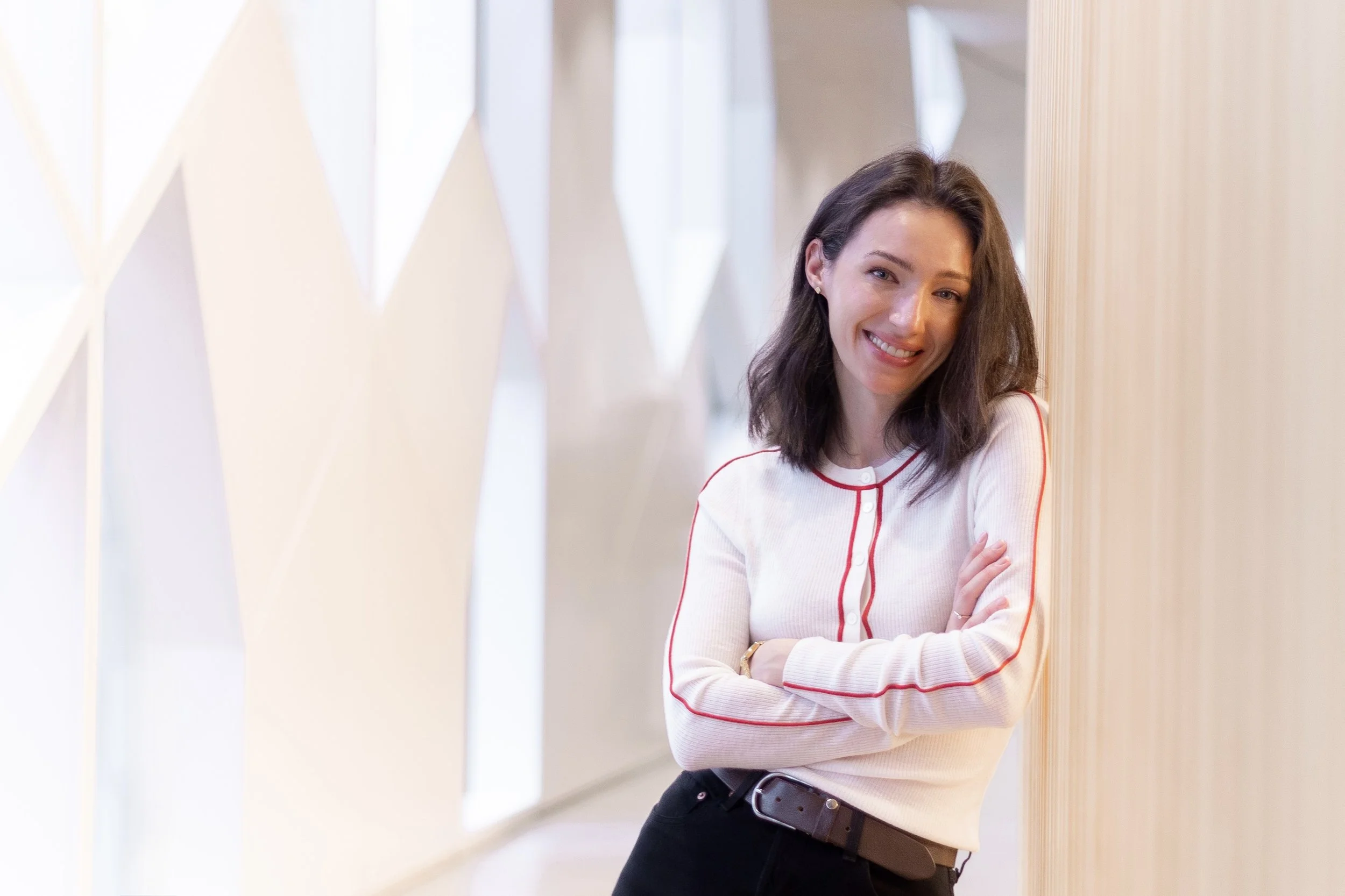 A woman with shoulder-length dark brown hair and a big smile, leaning against a wooden panel, wearing a white long sleeve shirt with red piping details and black pants, in a well-lit modern indoor space.