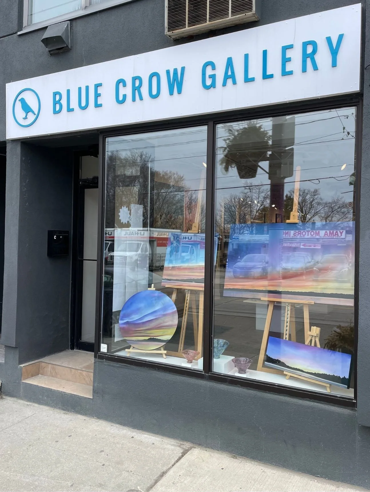 Storefront of Blue Crow Gallery with a white sign and blue lettering, showing paintings and pottery inside through large glass windows.