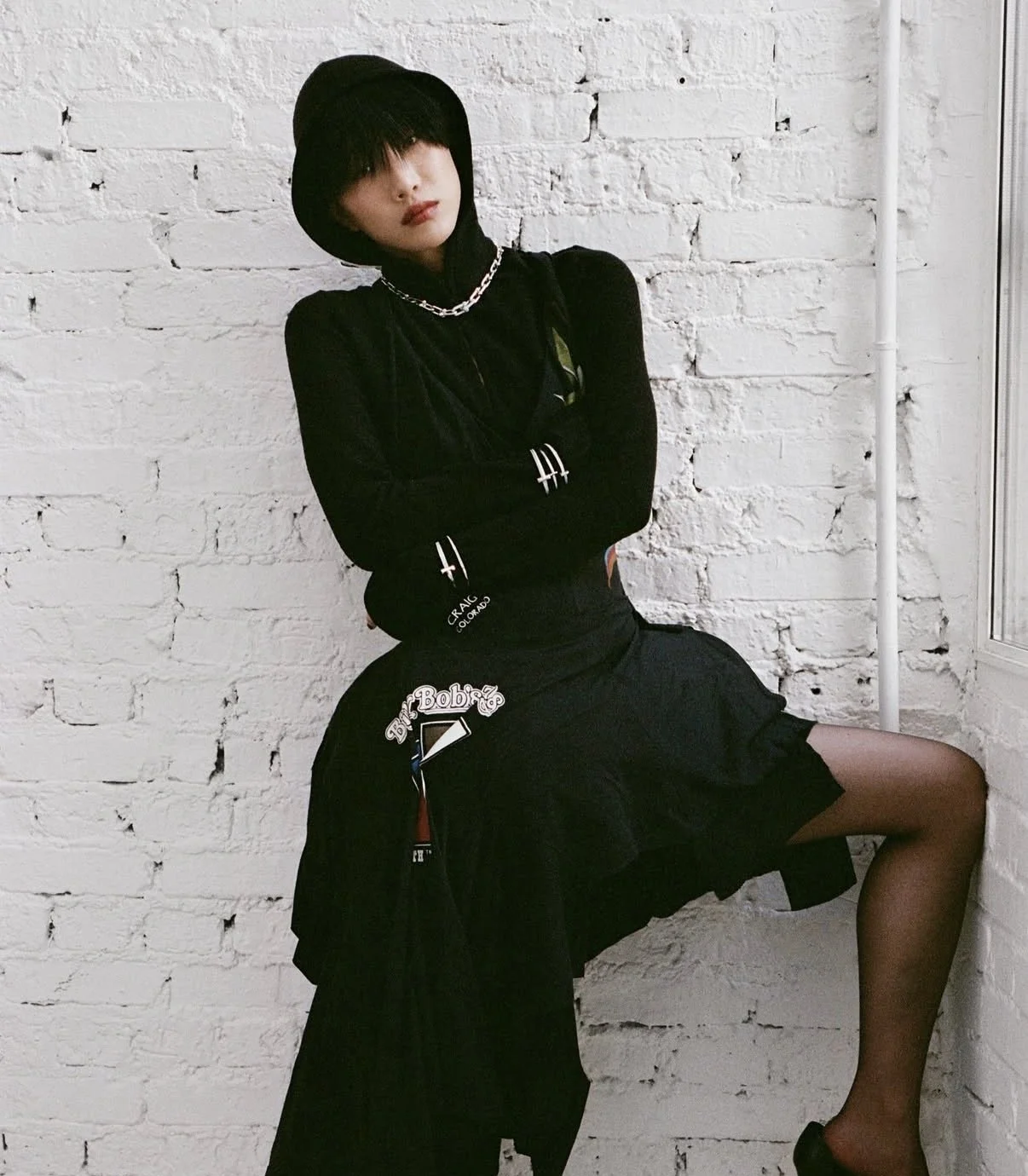 Woman with black hair and bangs wearing a black hooded top and black skirt, standing against white brick wall with arms crossed, one leg lifted and resting on the wall.