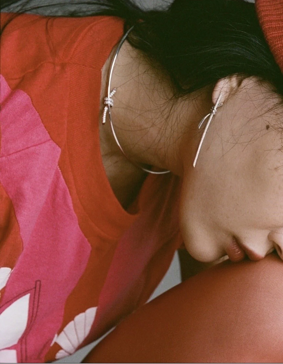 Close-up of a woman with long black hair wearing a red and pink hoodie, silver jewelry, and makeup, leaning forward with her face close to her knee.