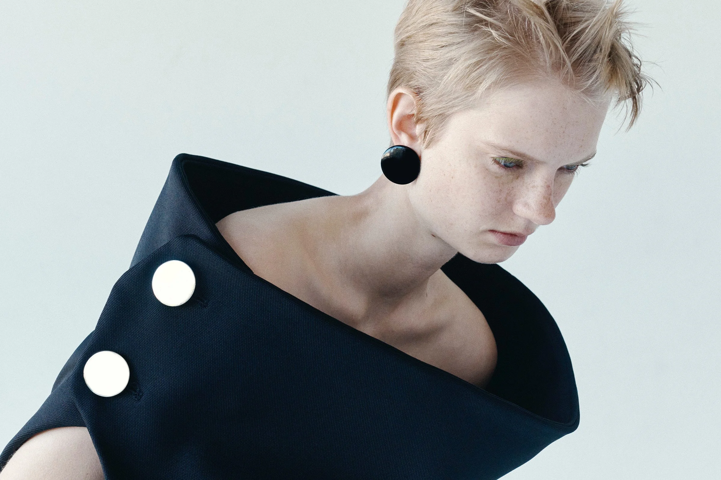Close-up of a woman with short blonde hair wearing a black dress with large white buttons and a black circular earring, looking down against a plain light background.