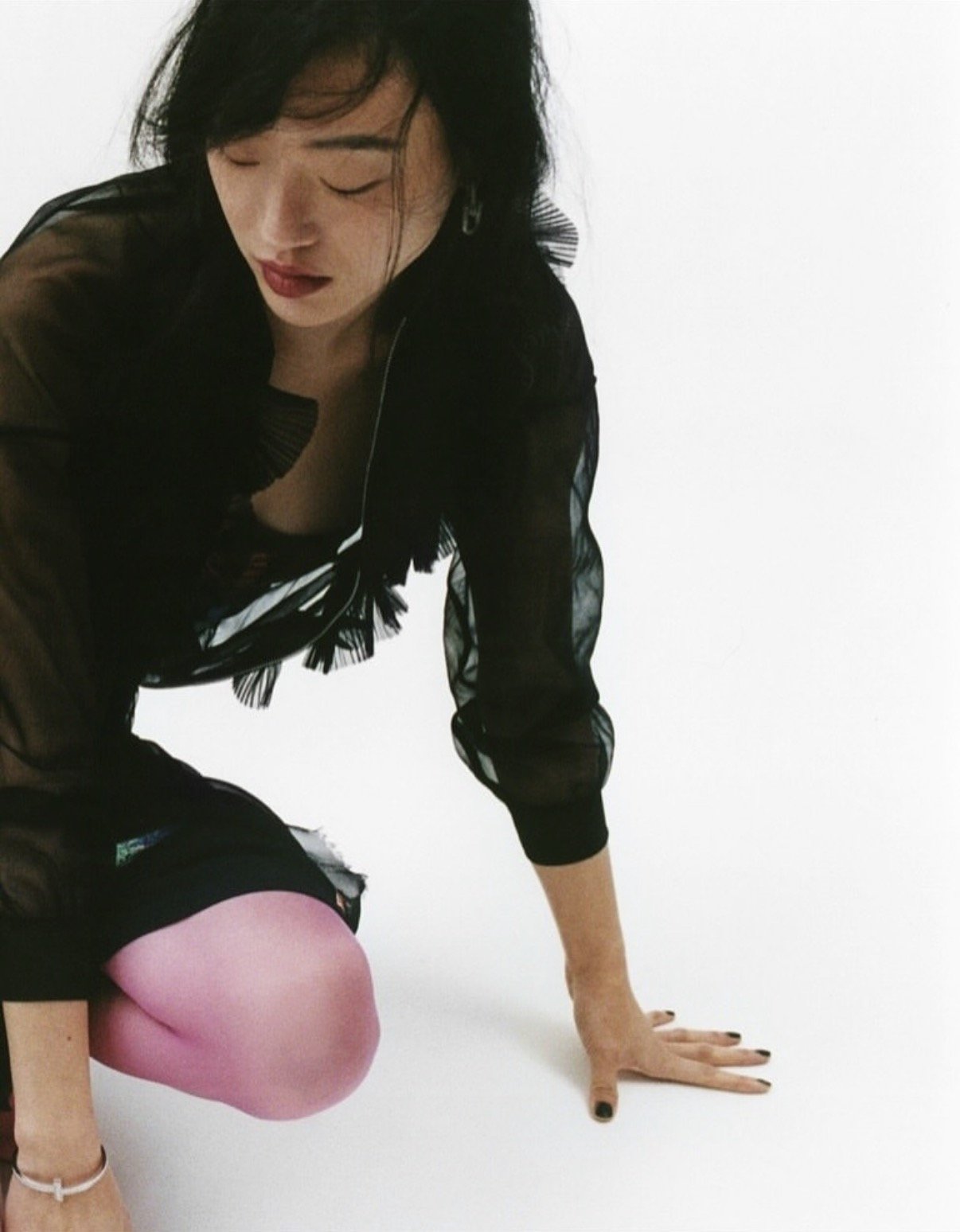 A woman with black hair and red lipstick is crouching on the floor, wearing a black sheer top, pink pants, and a white bracelet. She is looking down, with her eyes closed and head tilted slightly downward.