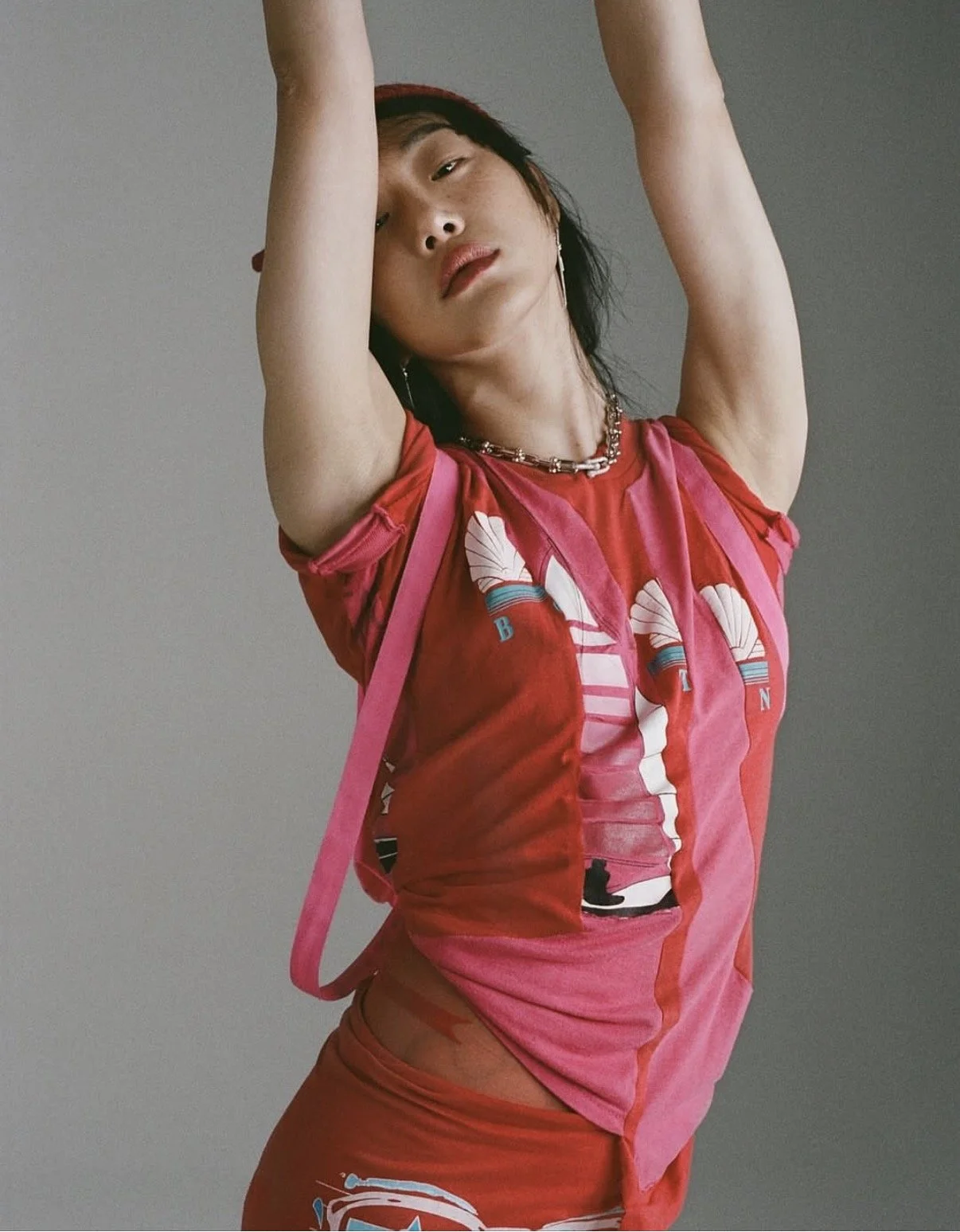 A person with short dark hair and light skin is posing with arms raised above their head, wearing a colorful red and pink graphic T-shirt, layered necklaces, and matching red pants, with a gray background.