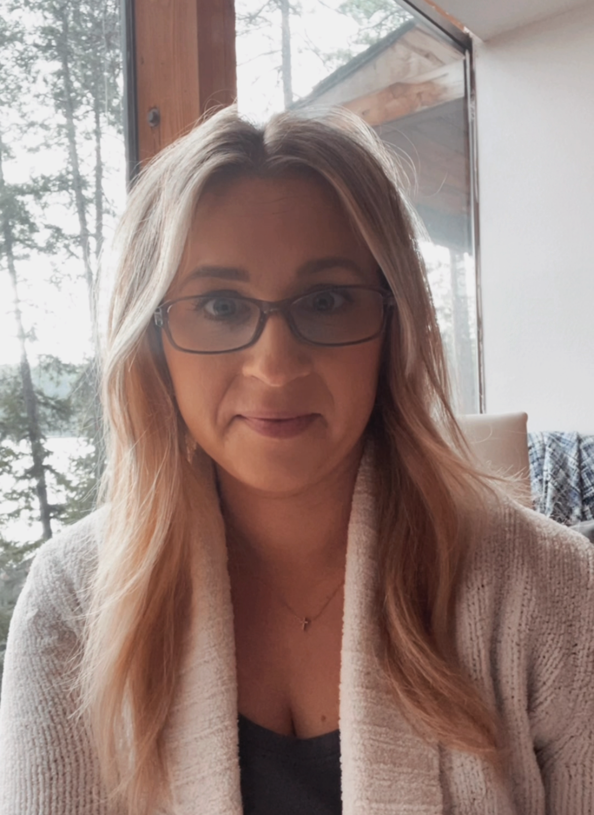 A woman with long, light brown hair, glasses, and wearing a beige sweater, sitting indoors near large windows showing trees outside.