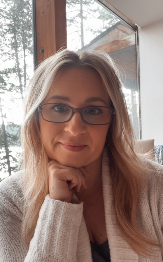 A woman with blonde hair and glasses resting her chin on her hand indoors near a window with trees outside.