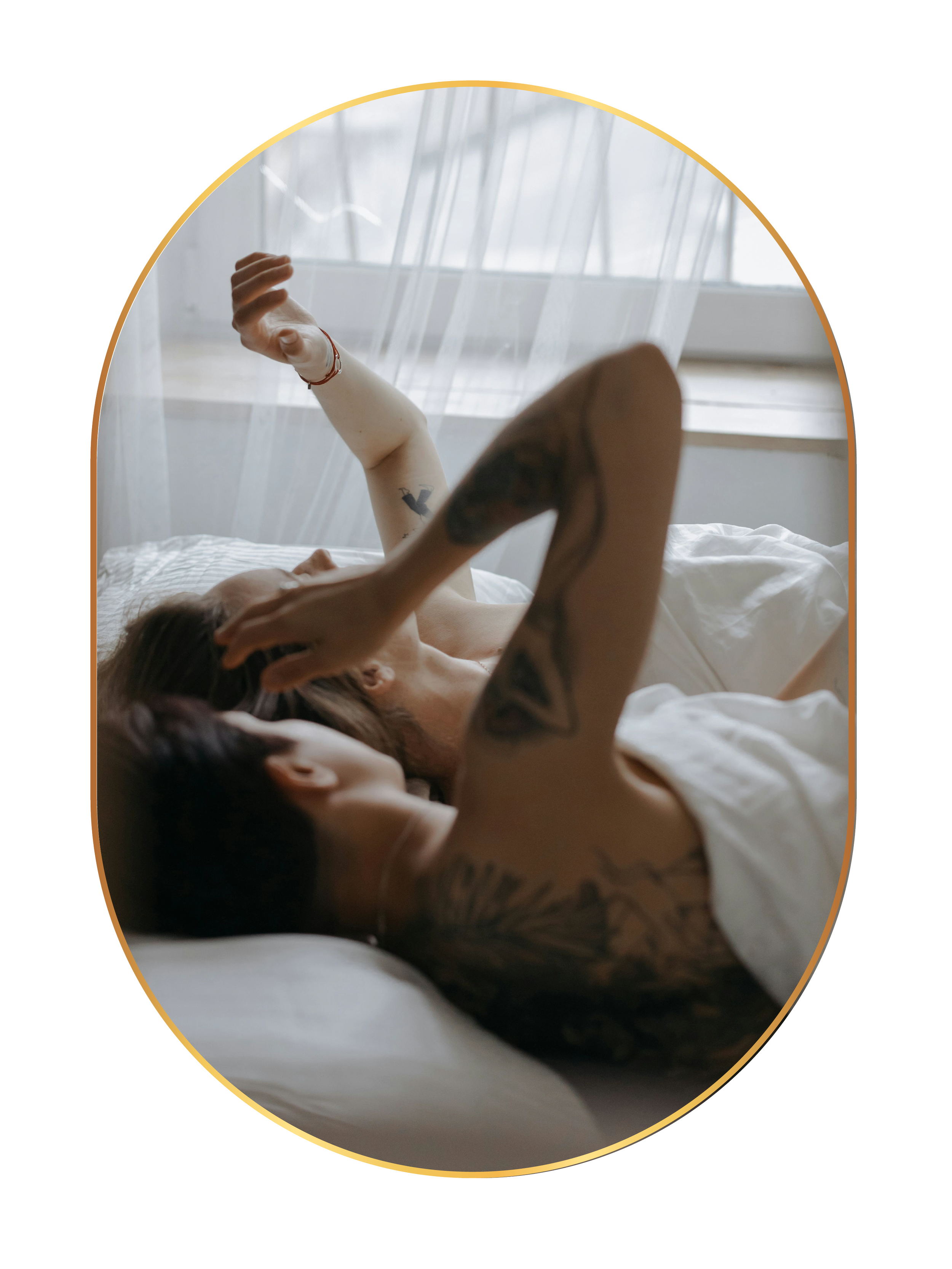 Two people with tattoos lying in bed, facing each other, with one touching the other's face, near a window with sheer curtains.
