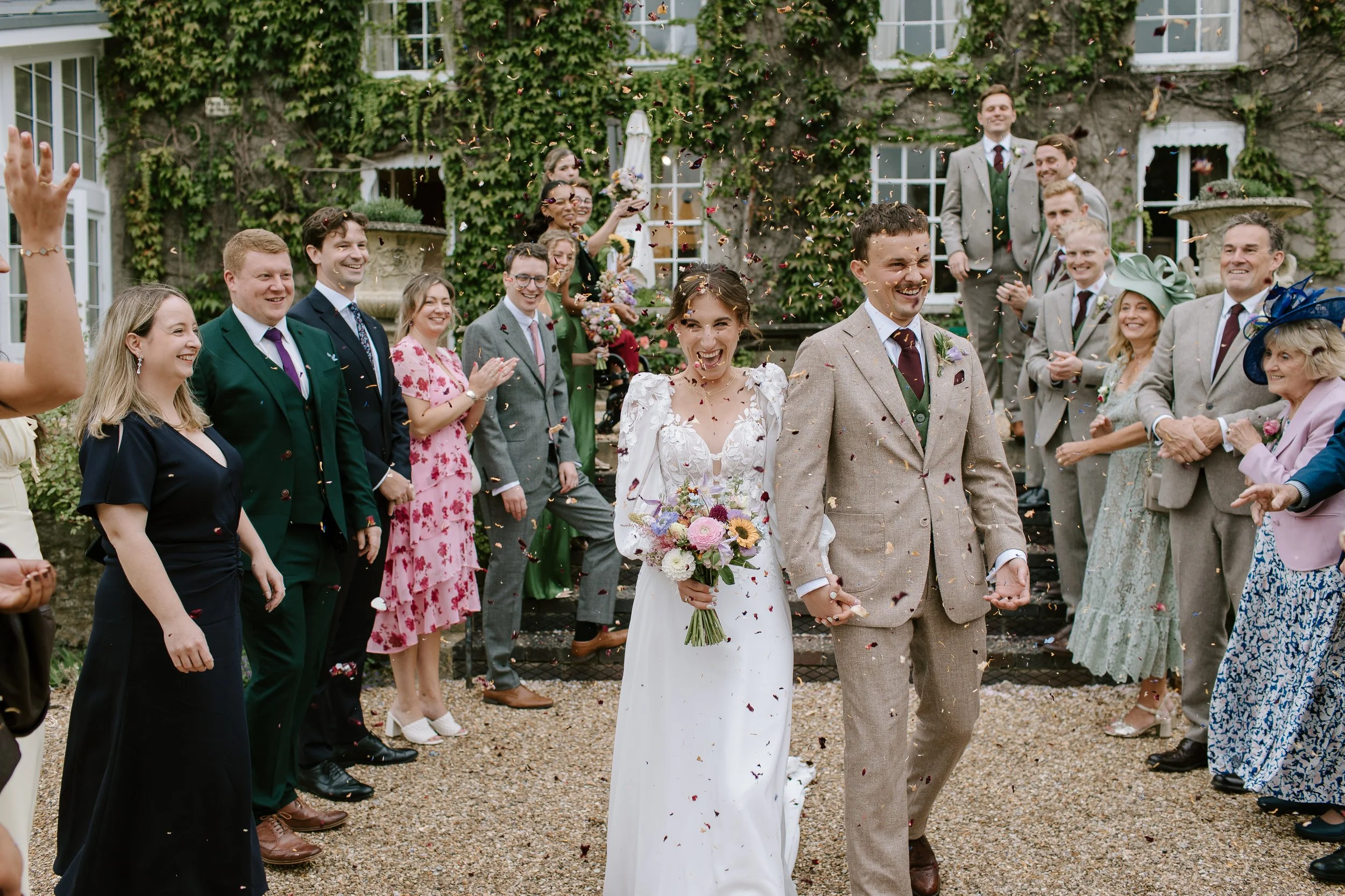 Bride and groom walking through confetti surrounded by guests outside a Somerset country house wedding venue.”