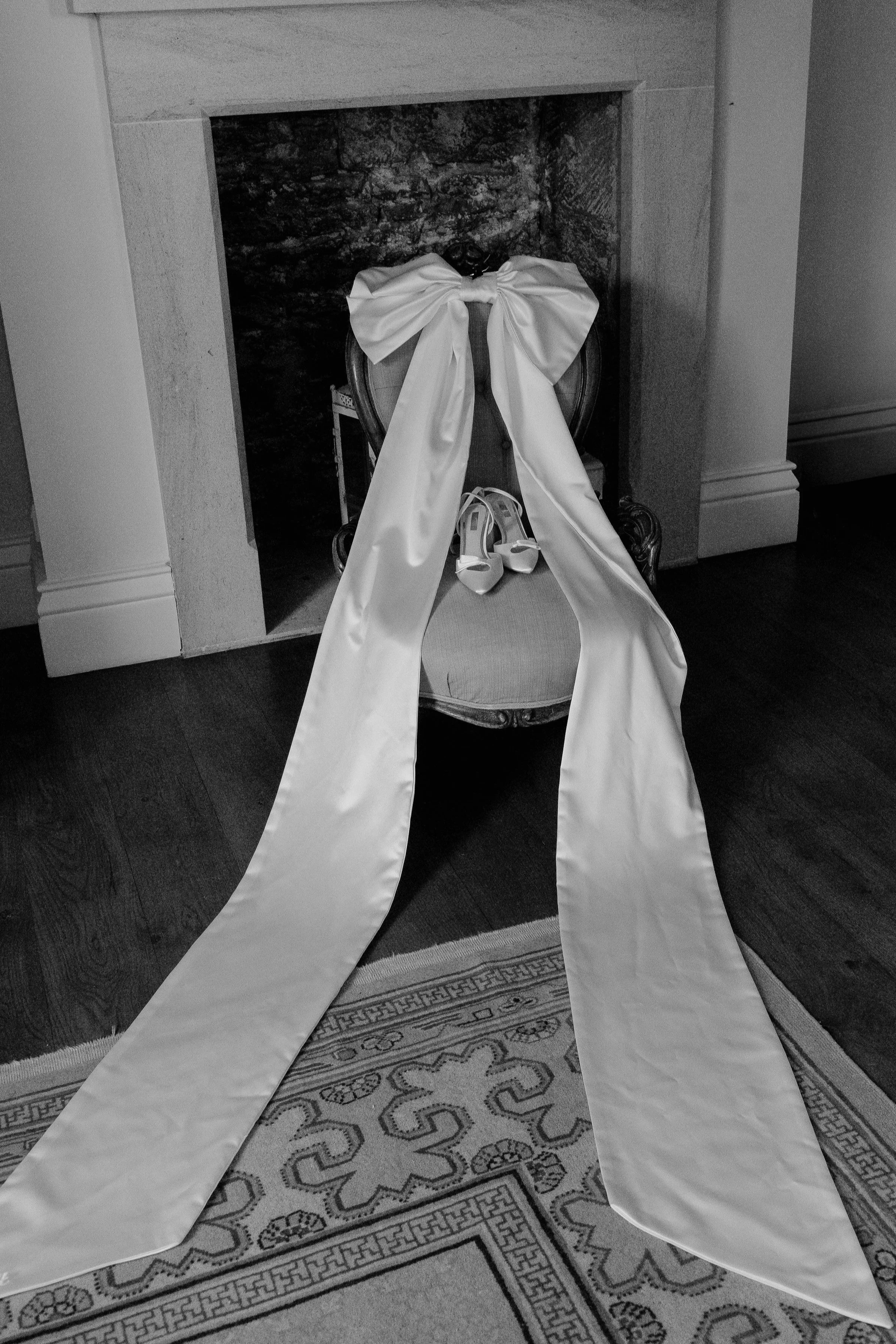 Black and white bridal detail photograph showing a large wedding dress bow and bridal shoes arranged on a chair indoors.