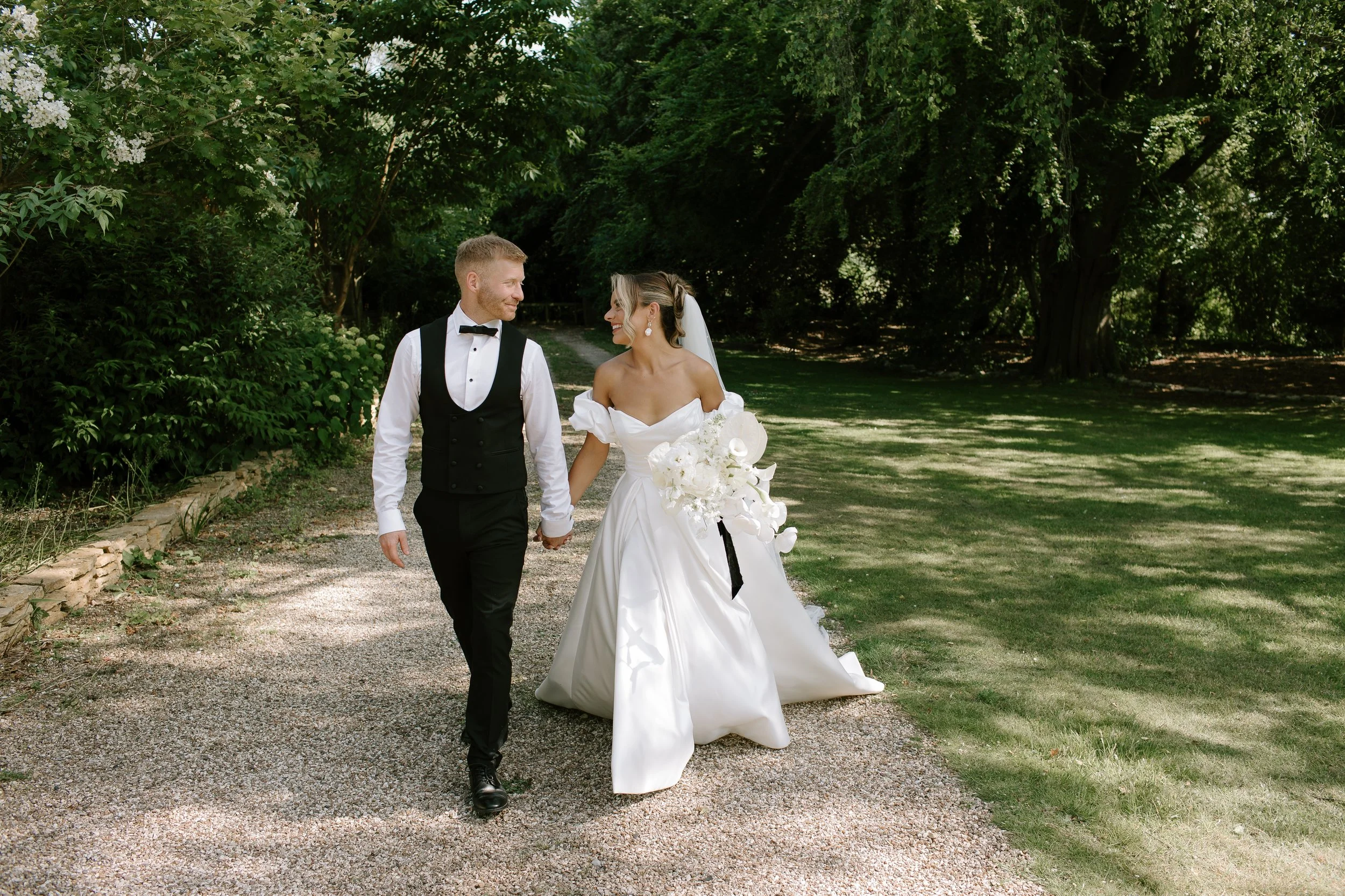 Bride and groom walking hand in hand along a garden path during a summer wedding at The Great Tythe Barn in Somerset.