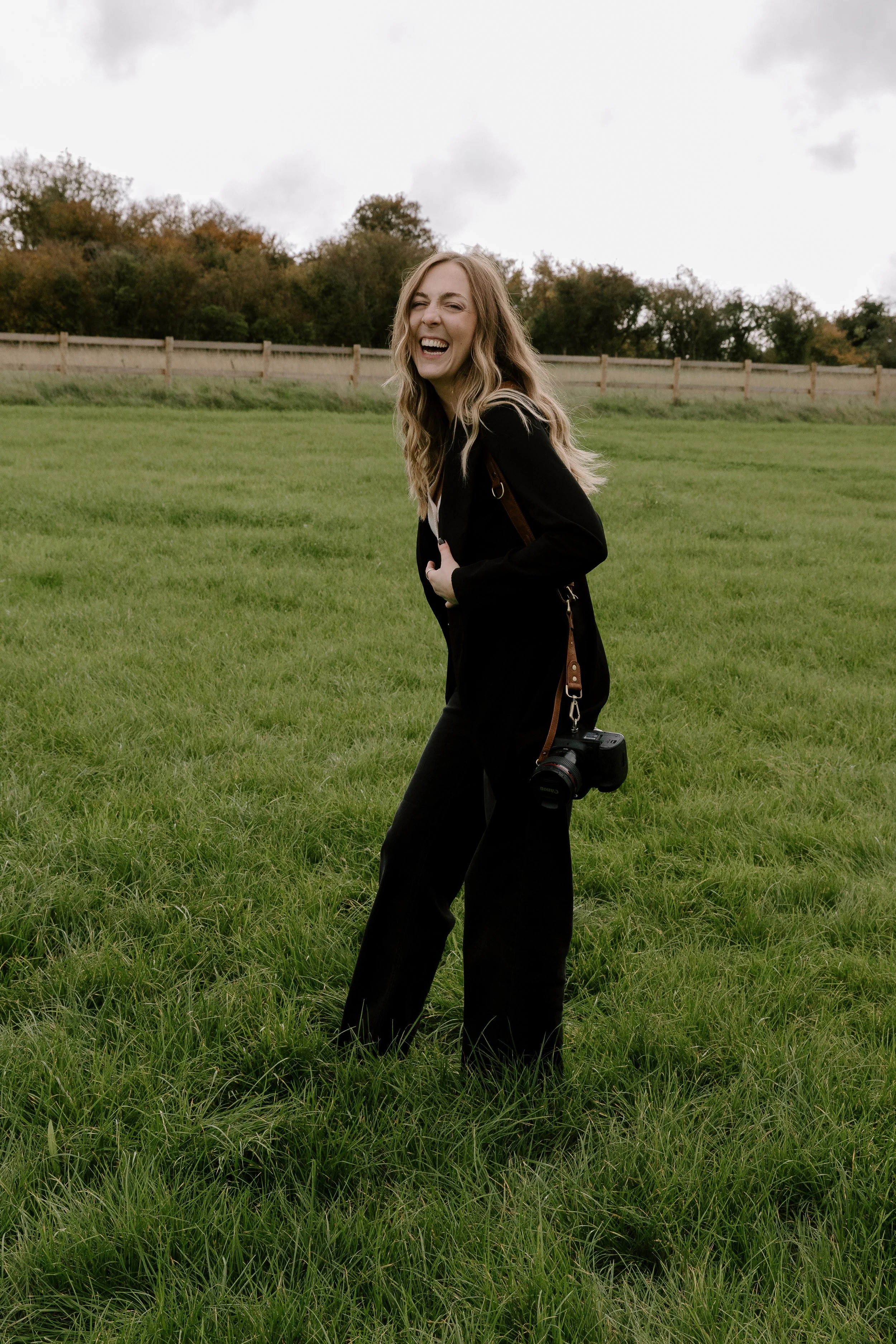 Wedding photographer walking through a grassy field in Somerset, smiling and holding a camera during an outdoor shoot.