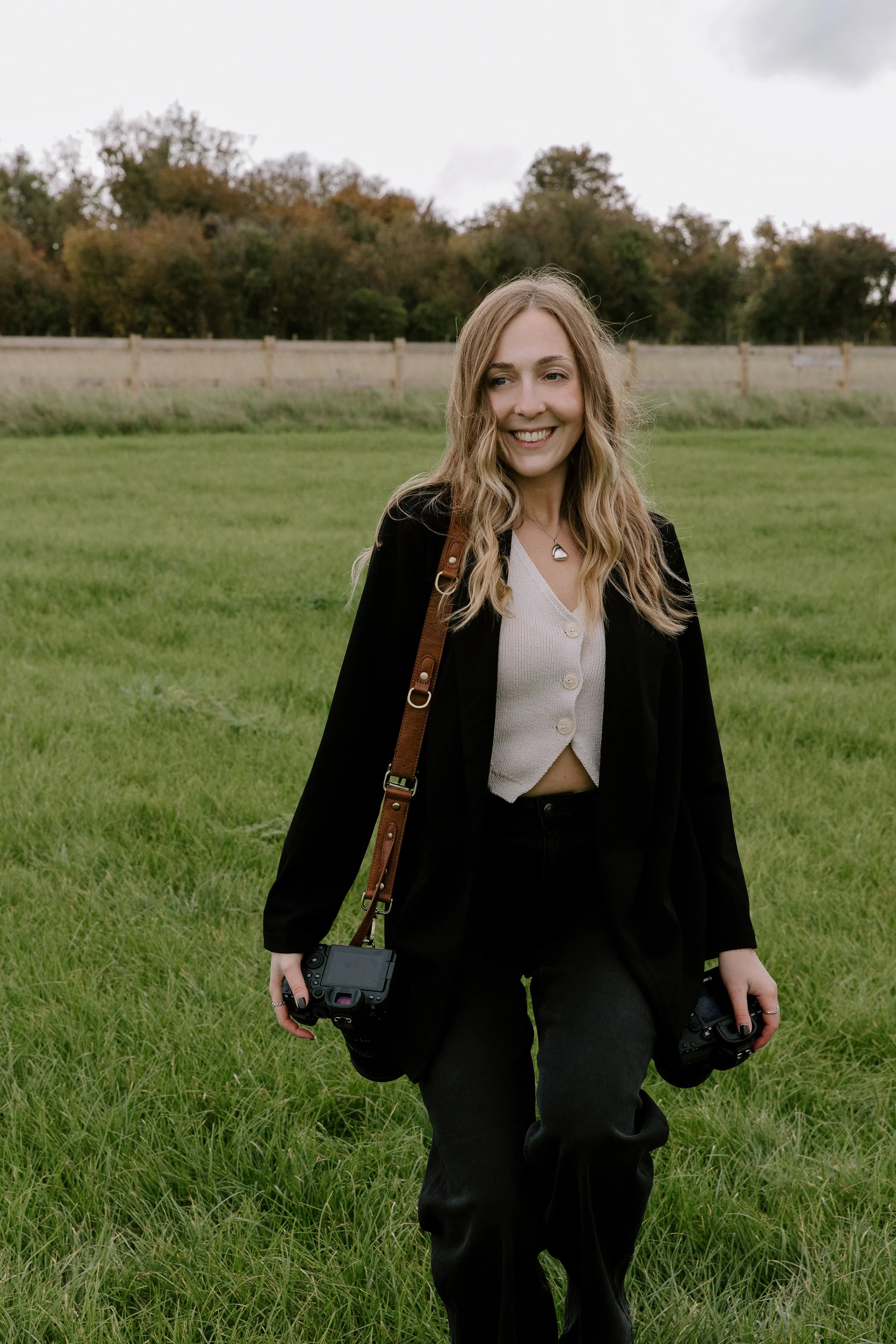 Portrait of Somerset wedding photographer Sian Hall walking through a field with camera gear, smiling naturally outdoors.