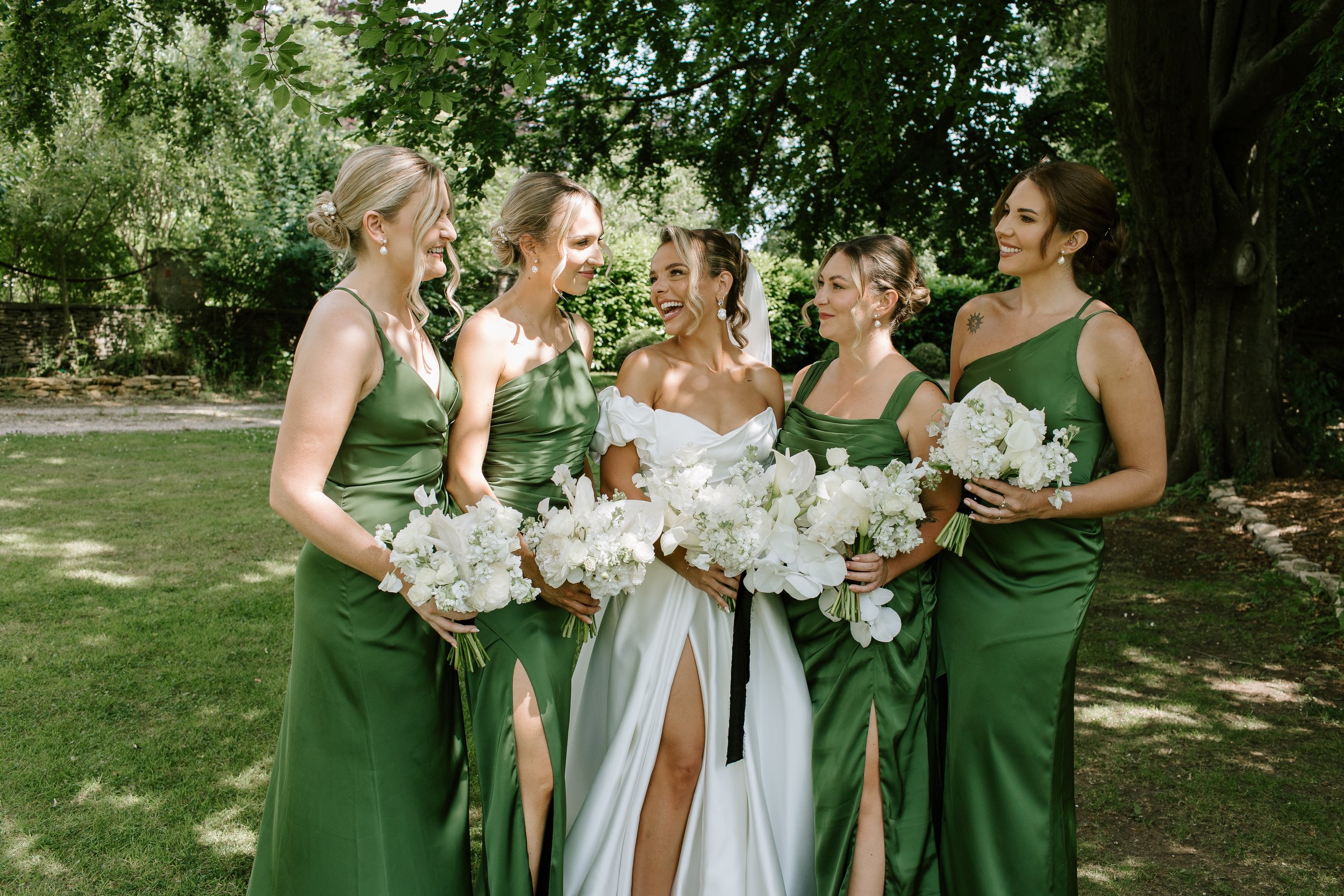 Romantic summer wedding portraits beneath the trees, with the bride surrounded by bridesmaids in elegant emerald green satin dresses and holding soft white bouquets. A timeless outdoor wedding moment filled with laughter, natural light, and modern br
