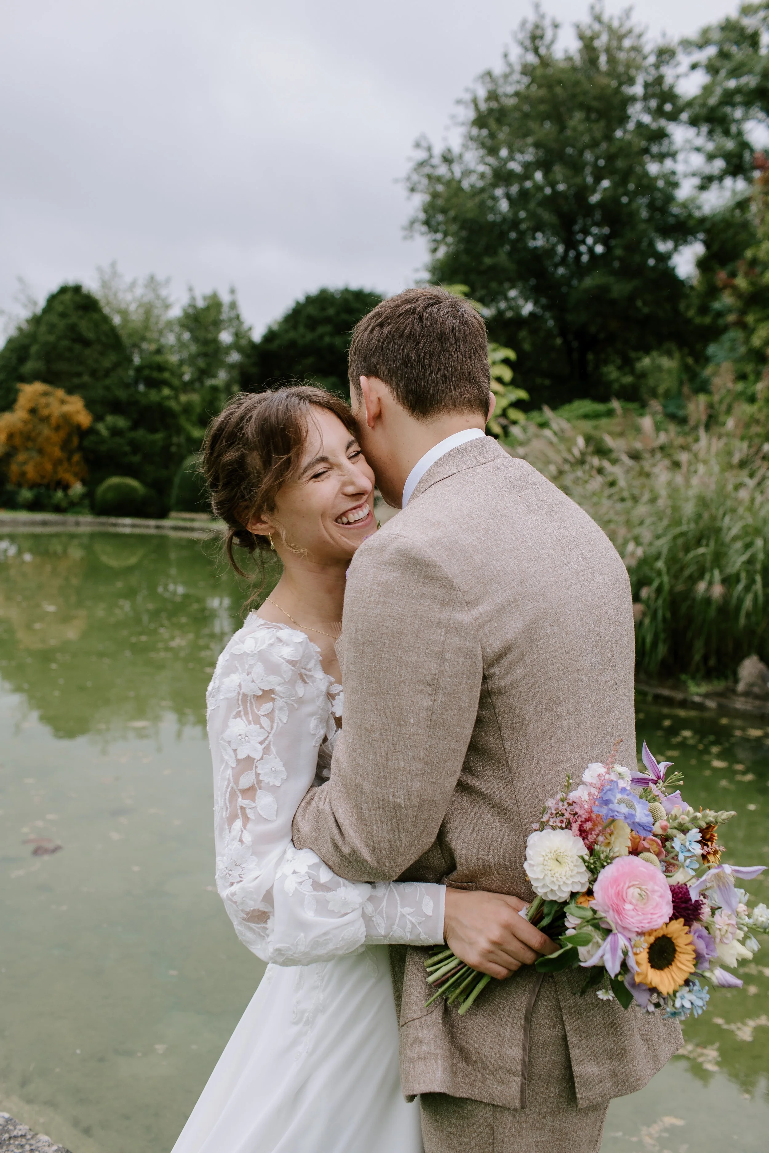 Bride and groom embracing beside a lake during relaxed wedding portraits at a Somerset country house wedding venue.