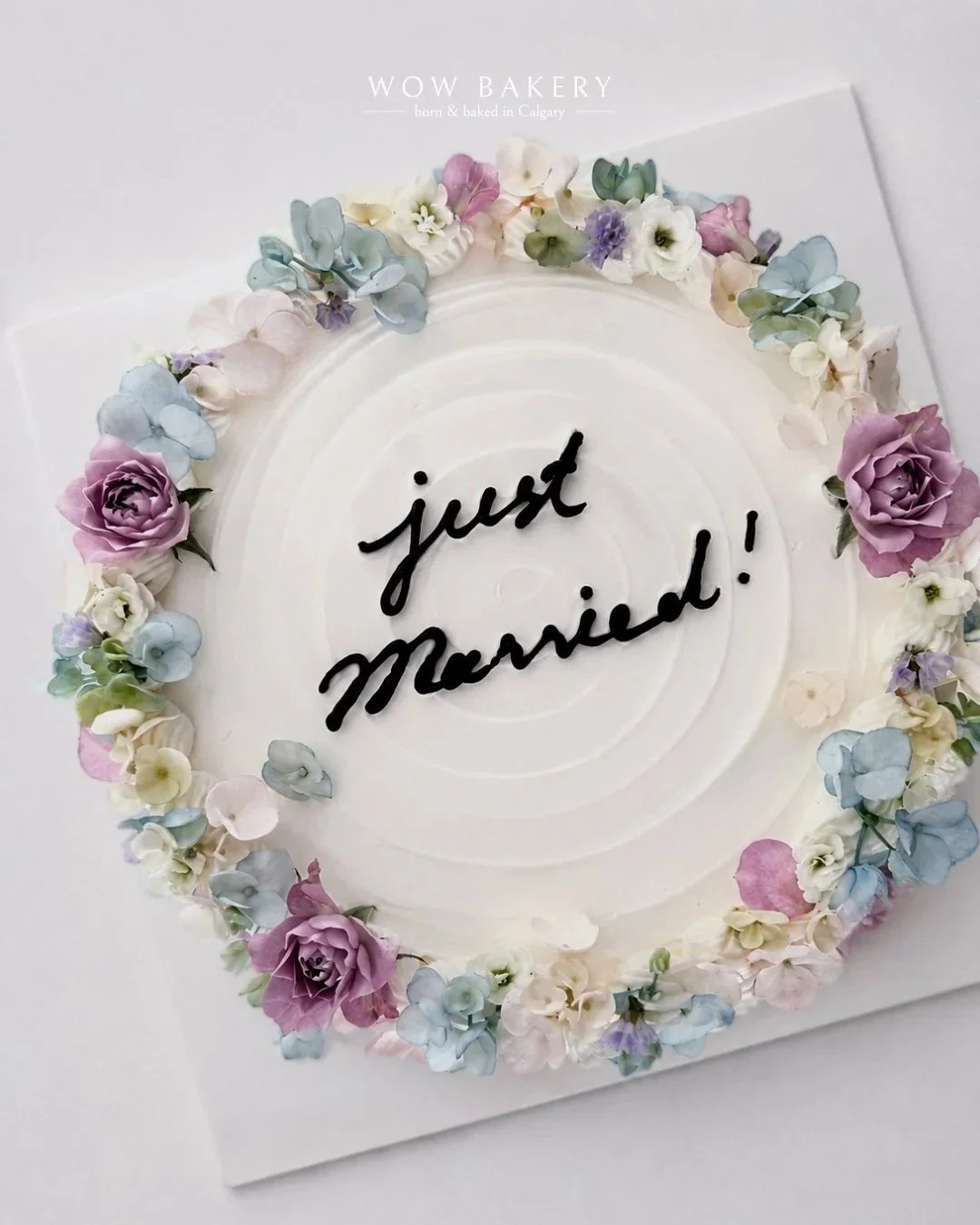Pastel floral “Just Married!” wedding cake with lavender, blue, and white flowers by WOW Bakery in Calgary.