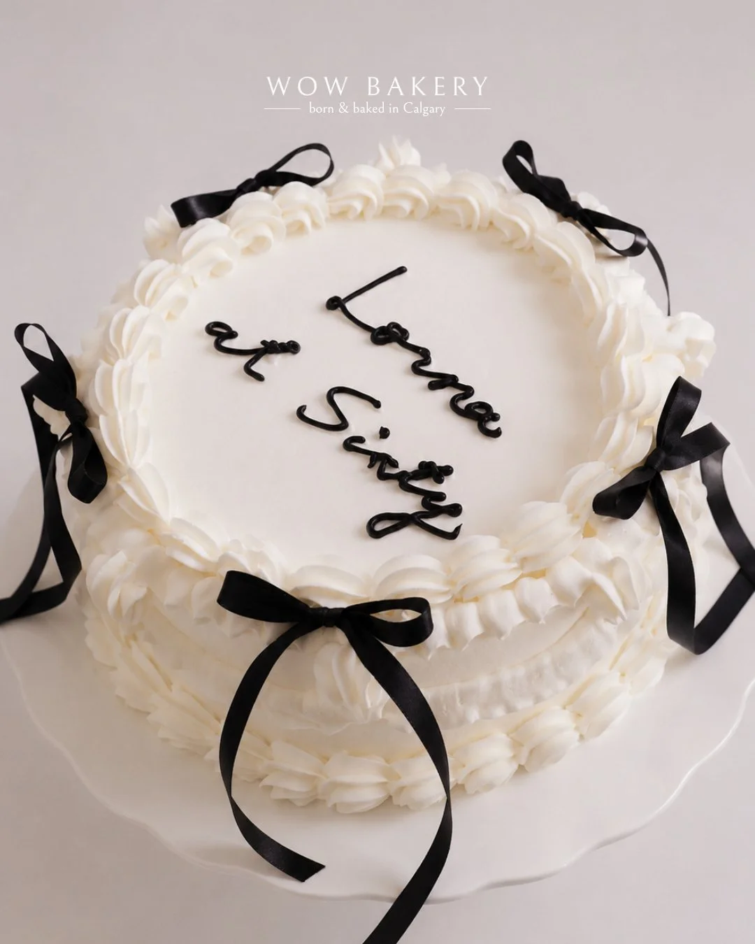 White vintage celebration cake with black ribbon bows and handwritten lettering, handcrafted by WOW Bakery in Calgary.