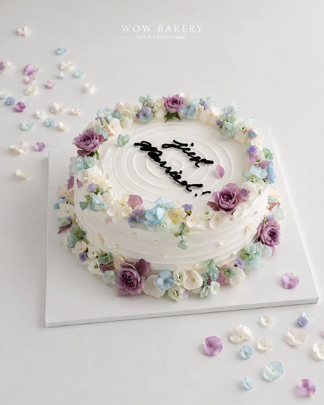 A white celebration cake decorated with pastel flowers and scattered petals, photographed on a clean white background.