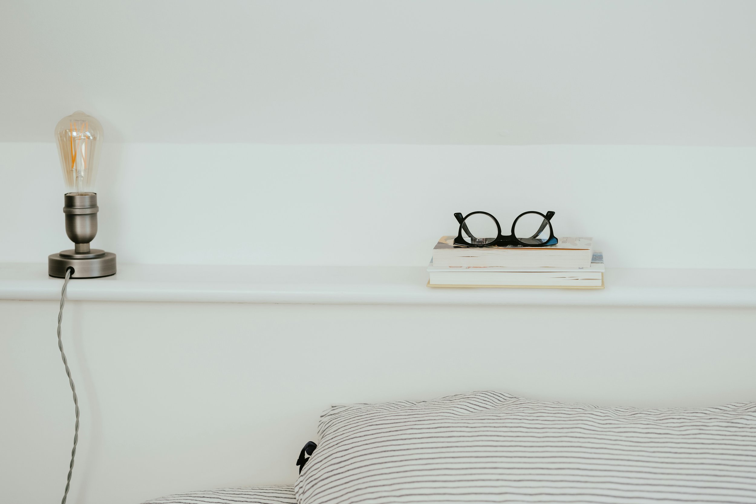 A minimalist bedroom scene with a white shelf. On the left side, there is a modern table lamp with an exposed Edison bulb and black base. On the right side, a stack of books with a pair of black round glasses resting on top. Part of a bed with a striped pillow is visible at the bottom of the image.