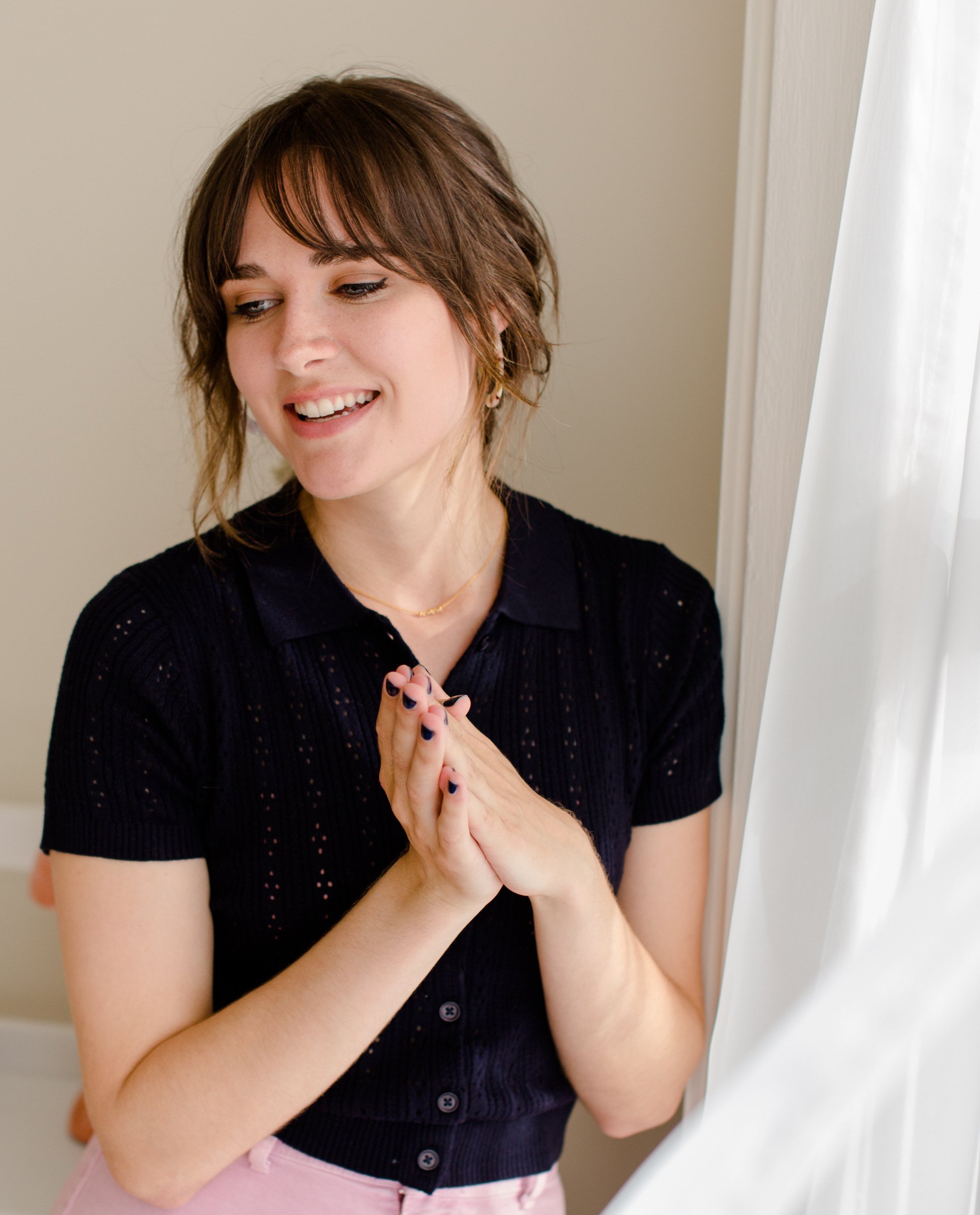 A smiling young woman with short brown hair and a black shirt standing near a window with white curtains.