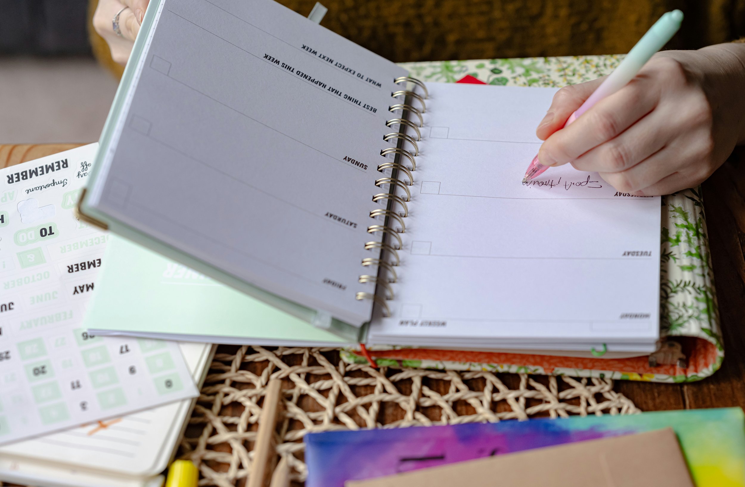 A person is writing in a spiral-bound planner or journal with a pink pen. The planner is open to a weekly layout with spaces for each day, and some text is visible on the right page. The planner is resting on a colorful, patterned cover. There are other papers, a calendar, and a woven placemat on the table.