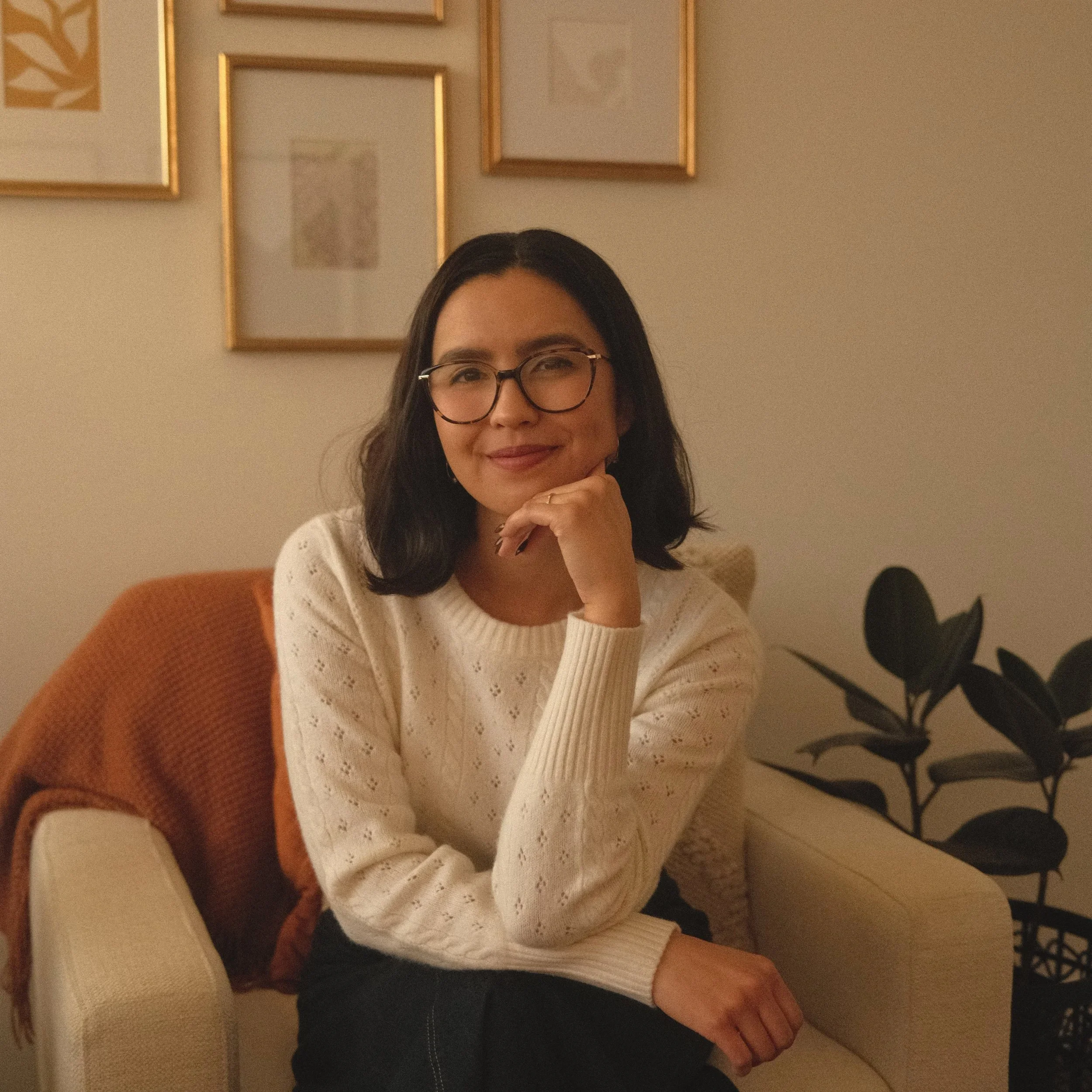 A woman with dark shoulder-length hair, glasses, and a white knitted sweater, sitting on a cream-colored armchair with an orange blanket draped over the back, in a cozy room with framed artwork on the wall and a leafy plant nearby.