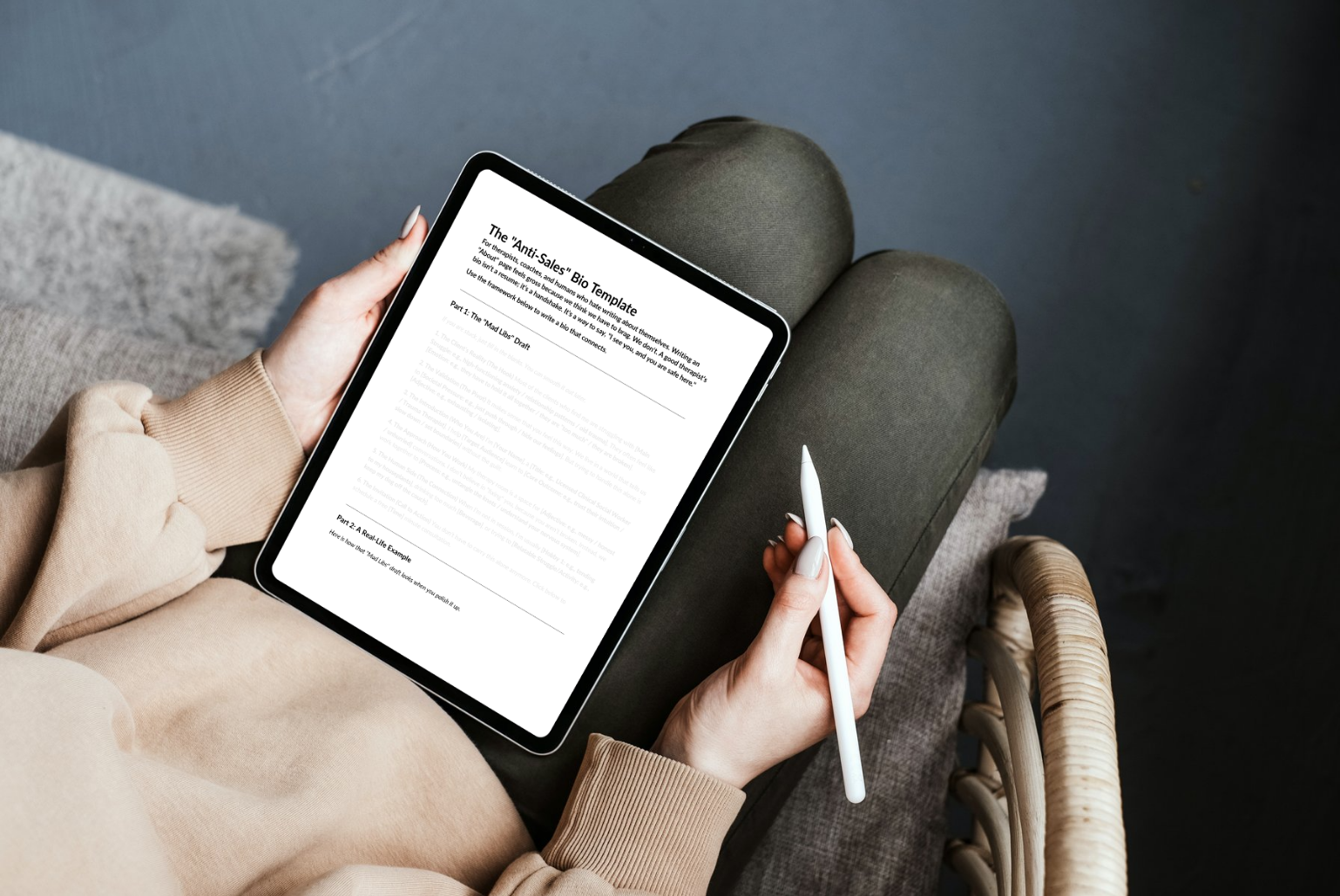 Person holding a digital tablet with text on the screen, sitting on a chair in a beige sweatshirt, with a stylus in hand, on a gray cushion and dark flooring.