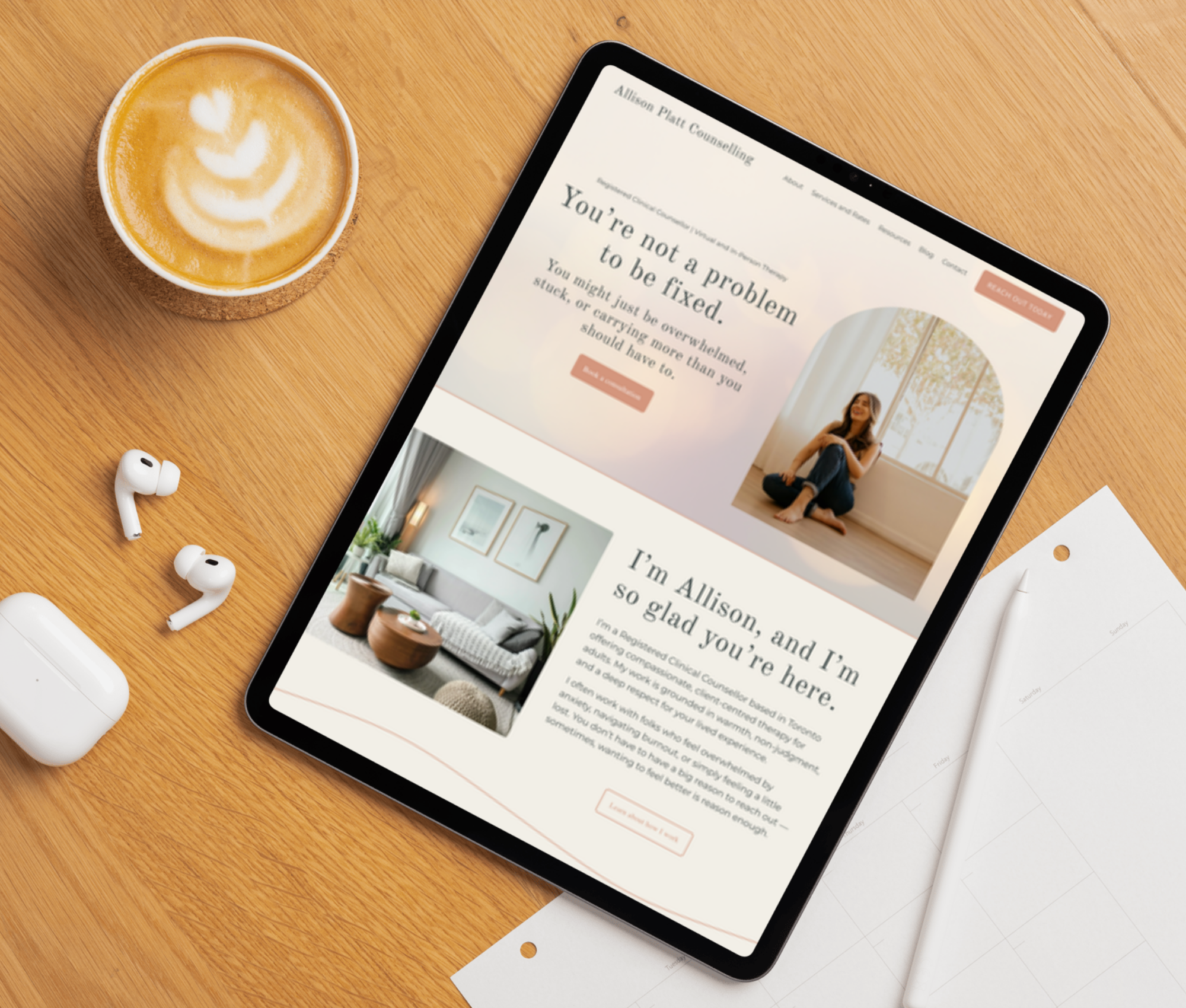A tablet displaying a website on counseling, placed on a wooden desk beside a cup of coffee with latte art, wireless earbuds, and sheets of paper.