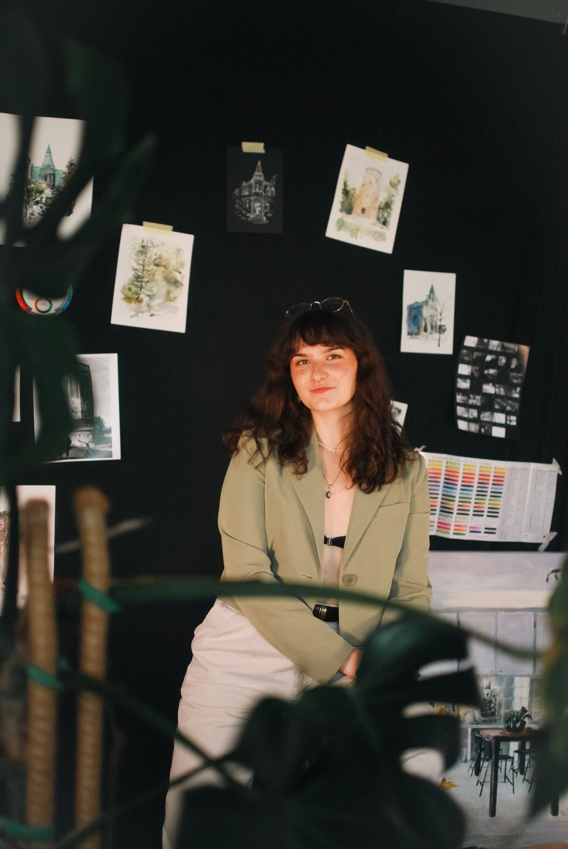 A woman with dark curly hair and glasses resting on her head, posing indoors with a relaxed smile, in front of a black wall decorated with various watercolor and black-and-white photographs and artwork.