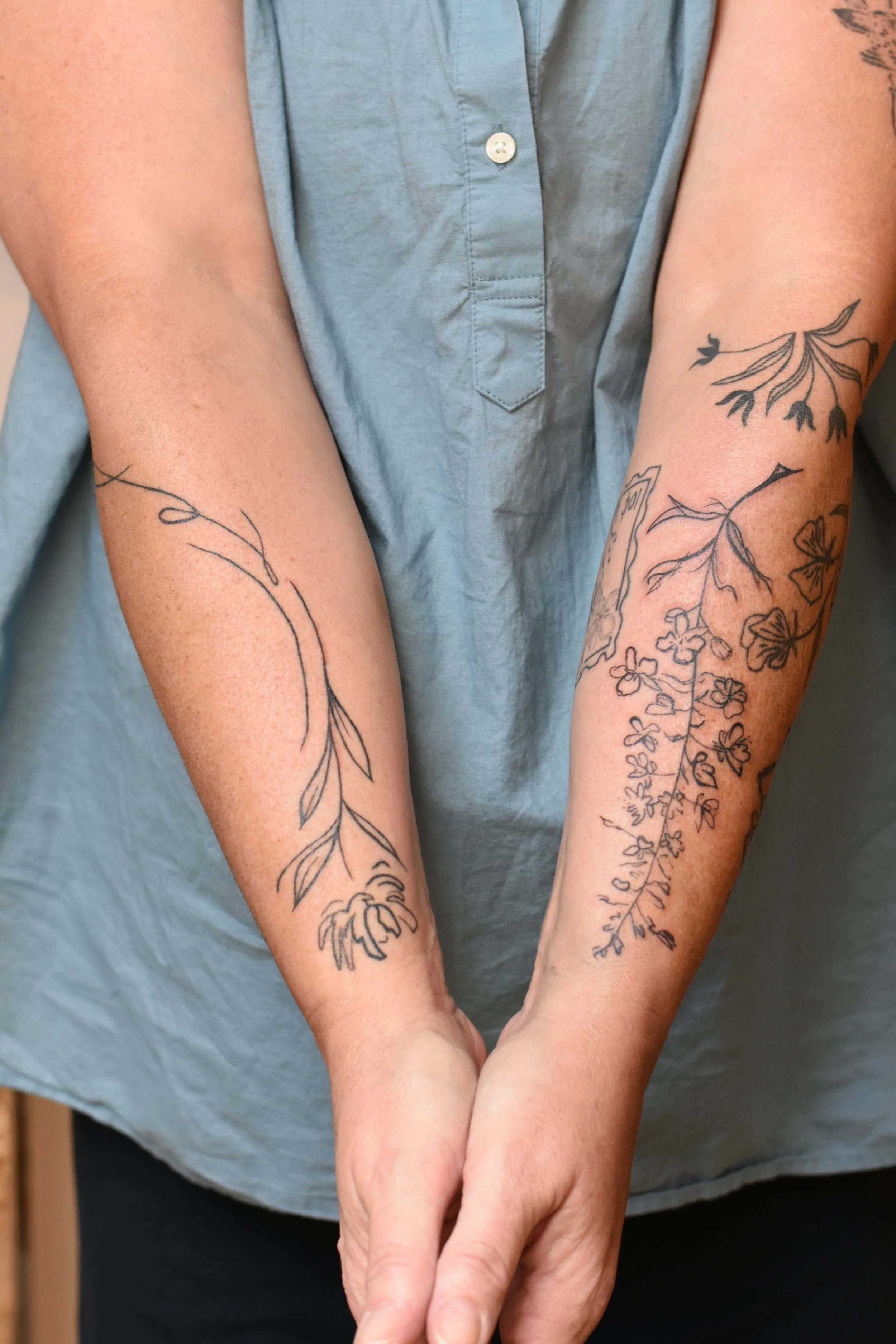 Person with two floral tattoos on their forearms, wearing a light blue sleeveless button-up shirt.