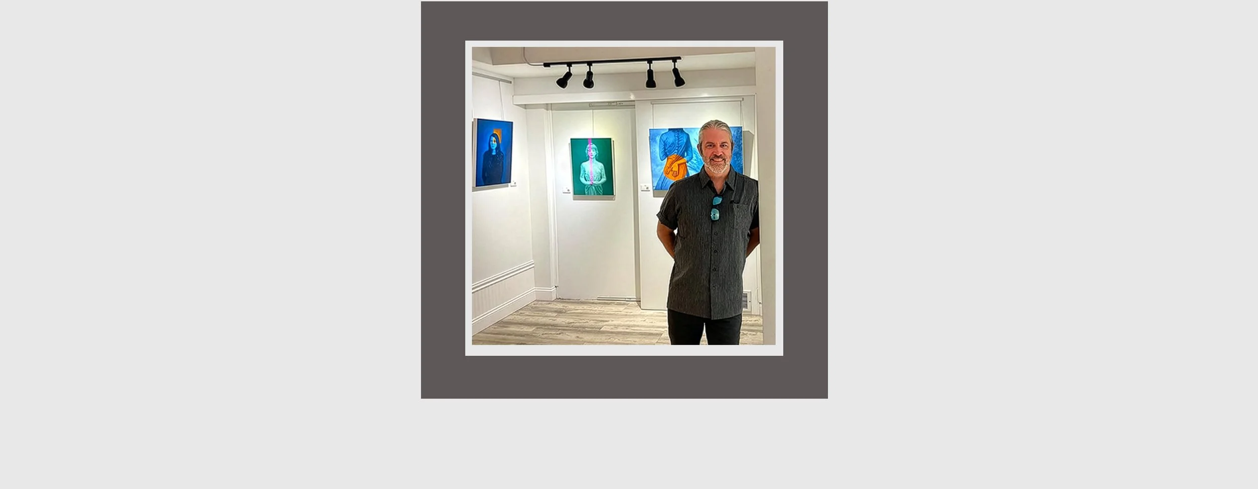 A man with gray hair and a beard smiling at an art gallery, wearing a dark shirt and sunglasses hanging from his collar, with colorful portraits hanging on the white walls behind him.