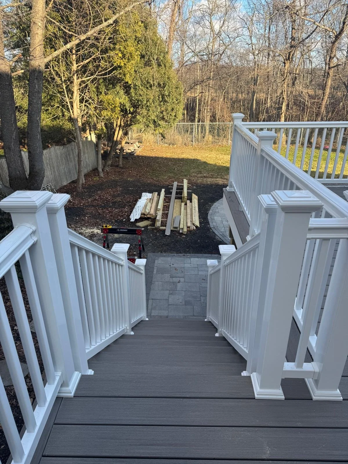 View from the top of a newly constructed white wooden deck stairs leading to a backyard with construction materials, trees, and a fence.