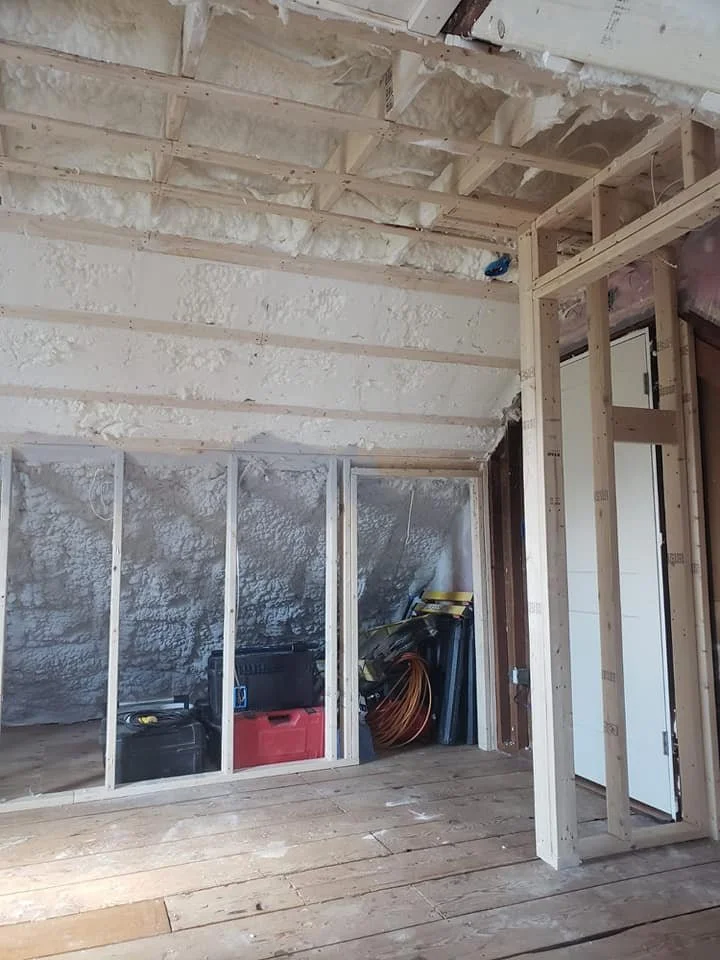 Interior of a construction site with unfinished walls and ceiling, showing wooden framing, insulation foam, and construction tools and materials on the floor.