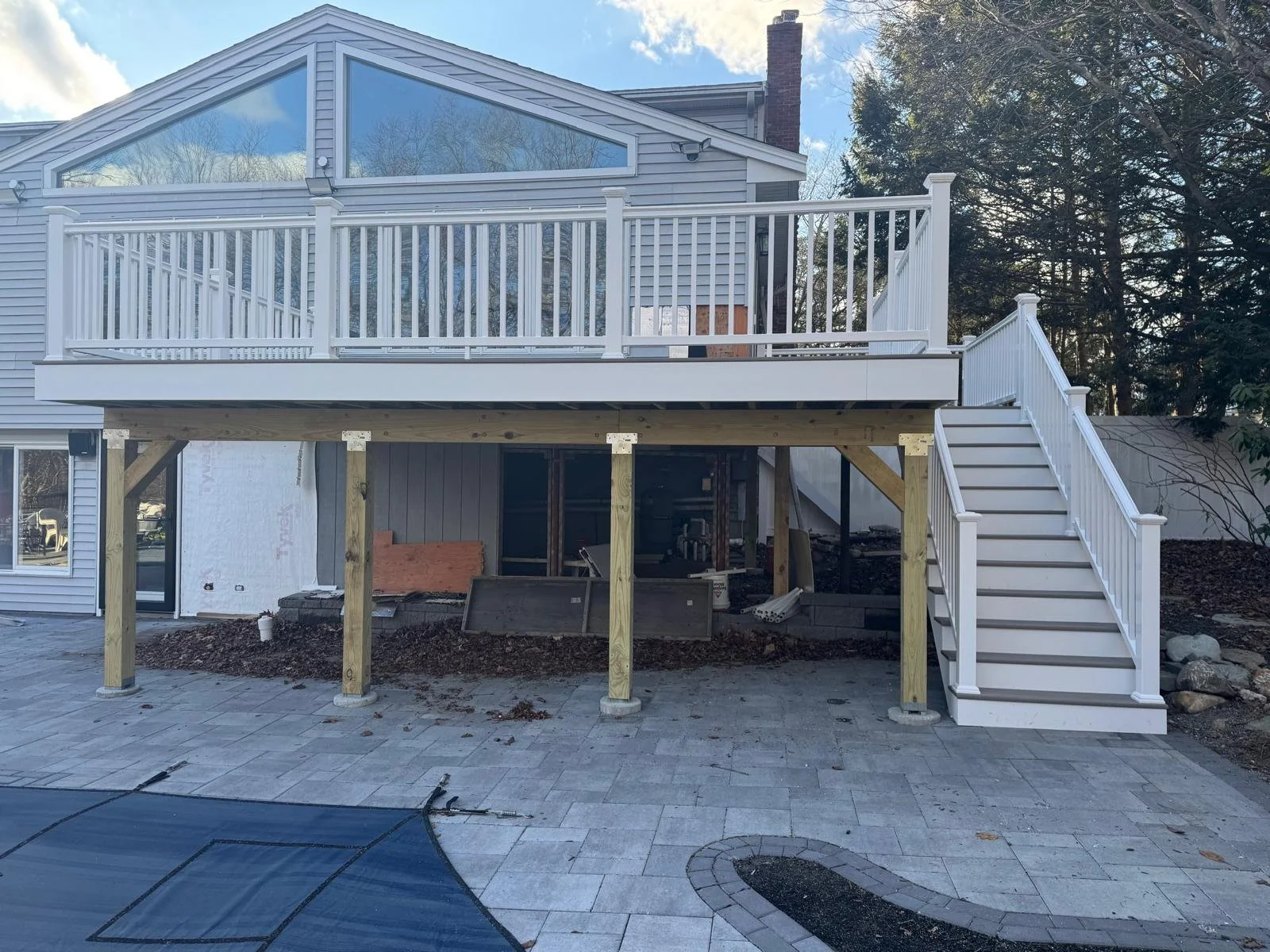 Backyard view of a house with a newly constructed elevated deck, white railing, and stairs leading down to a paved patio area, during daytime.
