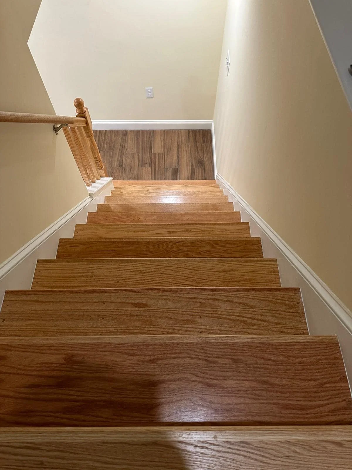 Wooden staircase viewed from the top, leading to a lower-level floor with hardwood flooring, beige walls, and electrical outlets.