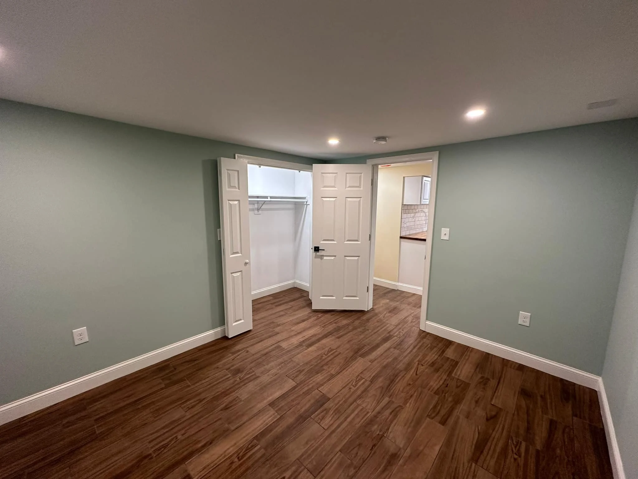 Empty room with hardwood floors, light green walls, and a closet with bi-fold doors. There is an open doorway leading to a hallway and a kitchen area.