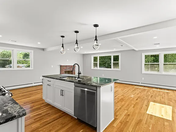 Modern open-concept kitchen with white cabinets, black granite countertops, a stainless steel dishwasher, and a kitchen island. Three hanging pendant lights are above the island. The space features hardwood floors and large windows allowing natural l