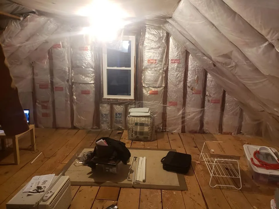 Attic room with insulation, a window, a small table, and various stored items.