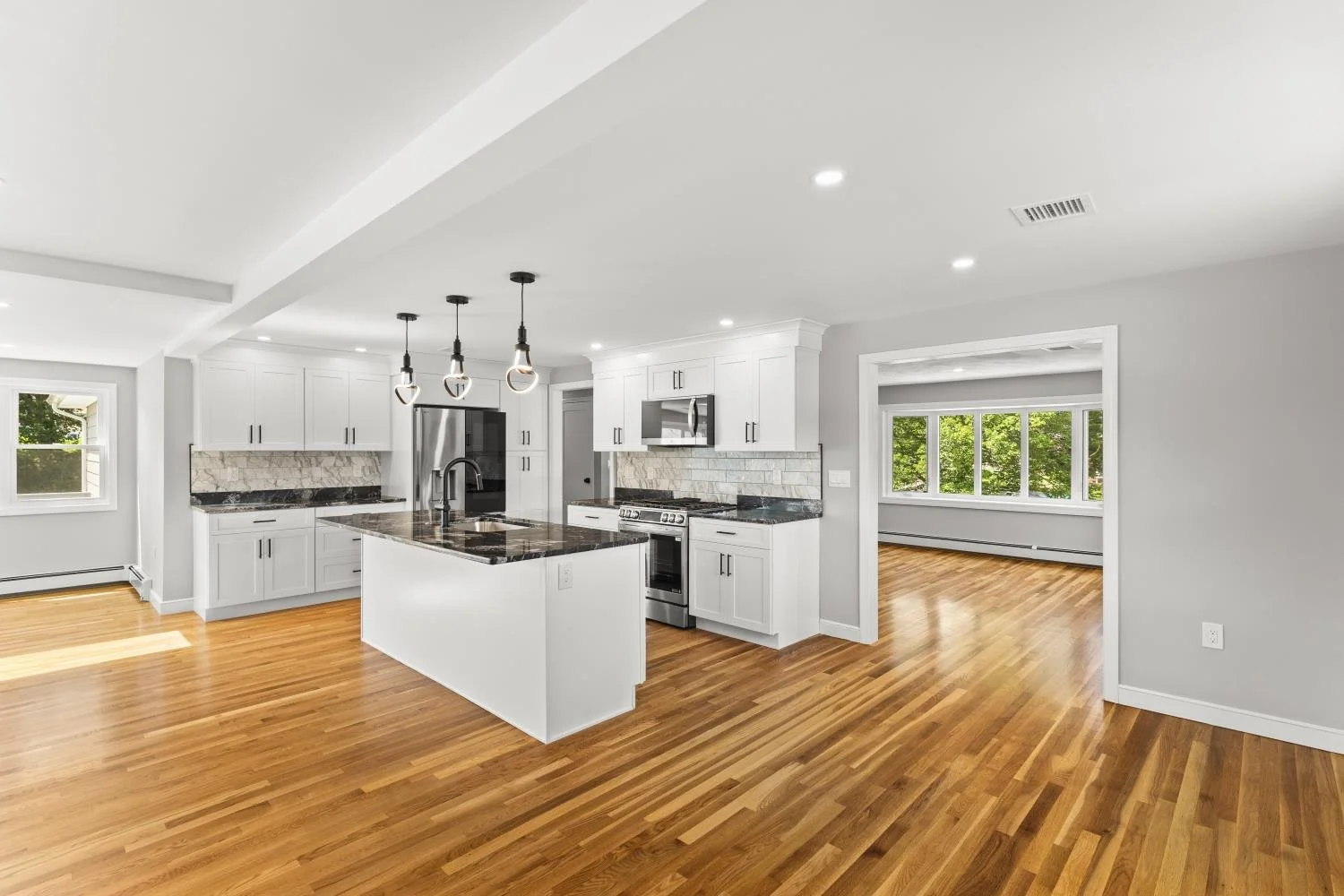 Modern kitchen with white cabinets, black countertops, stainless steel appliances, a central island, and hardwood floors, with adjacent bright dining area and large windows showing greenery outside.
