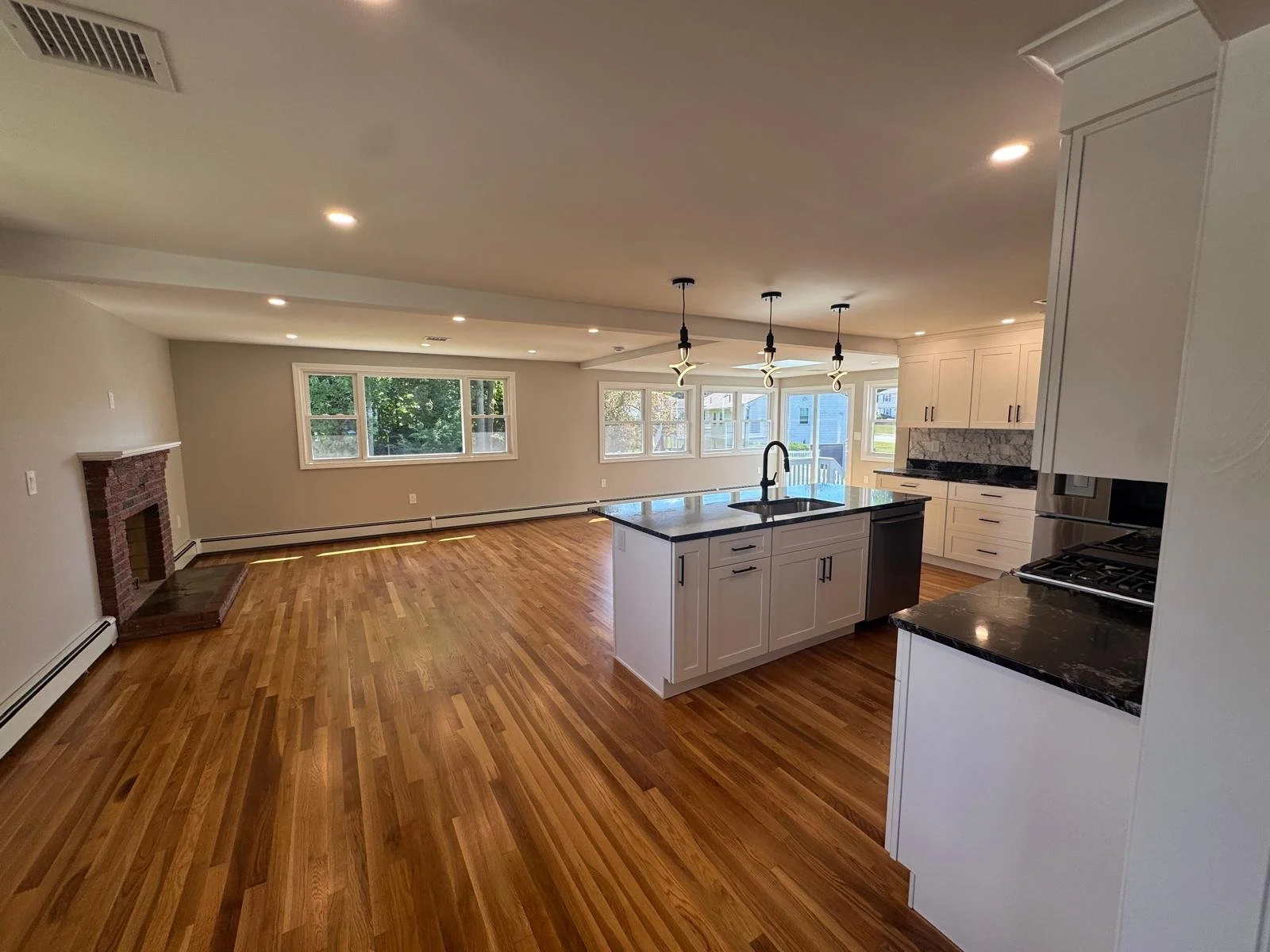 Open-concept living room and kitchen with hardwood floors, white cabinets, central island with sink, and large windows.
