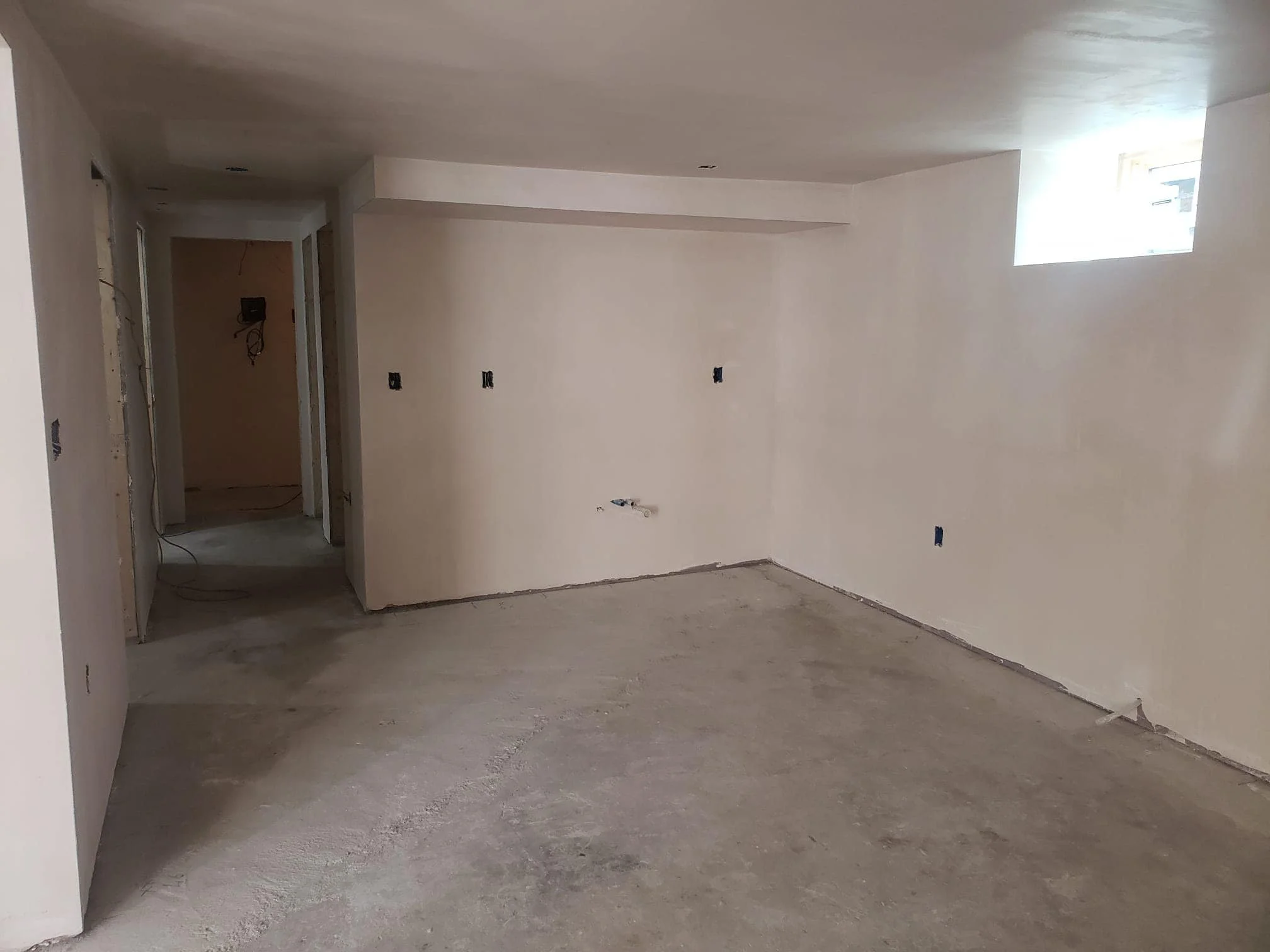 An unfinished room with drywall installed, electrical outlets without covers, and a concrete floor with visible marks and patches.