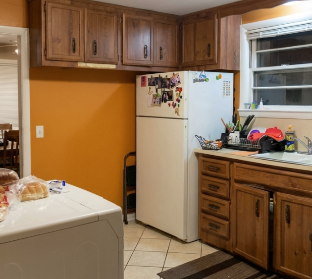 Kitchen with yellow wall, wooden cabinets, white refrigerator with photos and magnets, window above sink, dish rack with dishes and utensils.