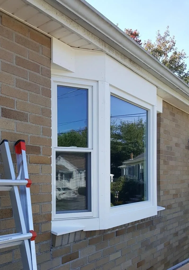 Close-up of a bay window in a brick house with a ladder nearby, showing the house's exterior wall, window trim, and roofline.