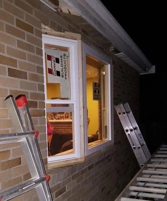 View of a brick house under construction with two ladders leaning against the wall and a large window installed, showing the interior.