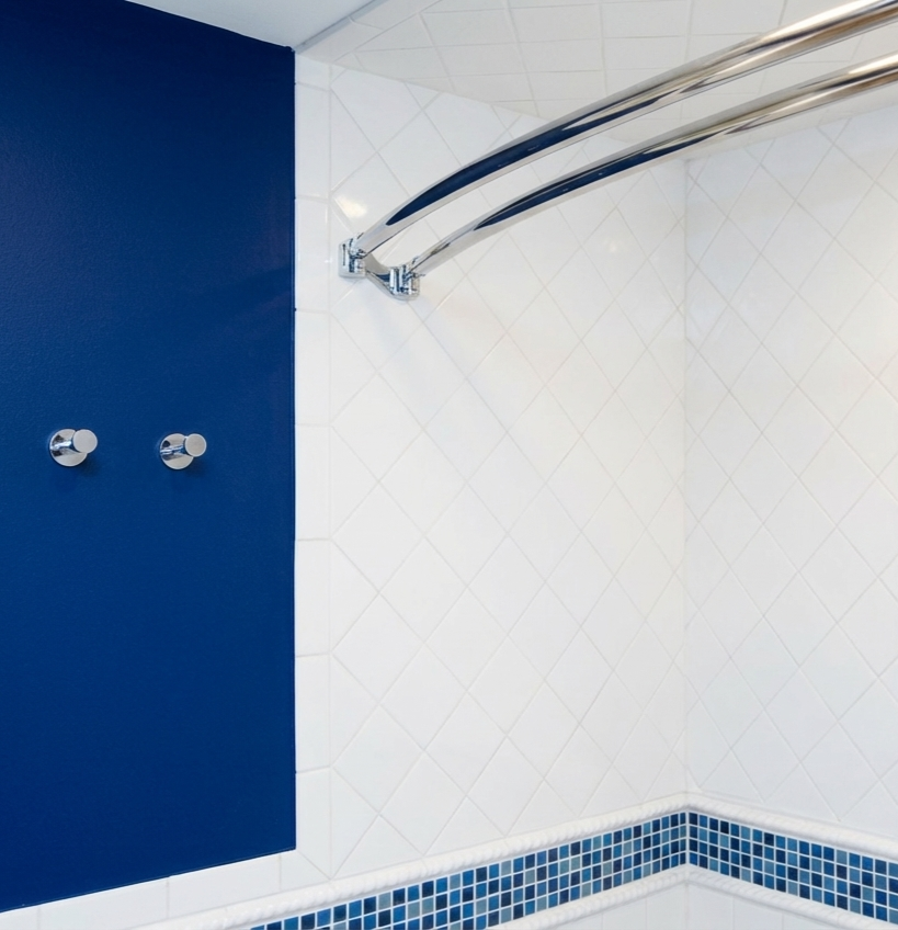 A shower with white tiled walls, a blue accent wall, and a row of small blue tile accents at the bottom. Two wall hooks are on the blue wall, and a curved chrome shower rod is mounted on the white tiles.