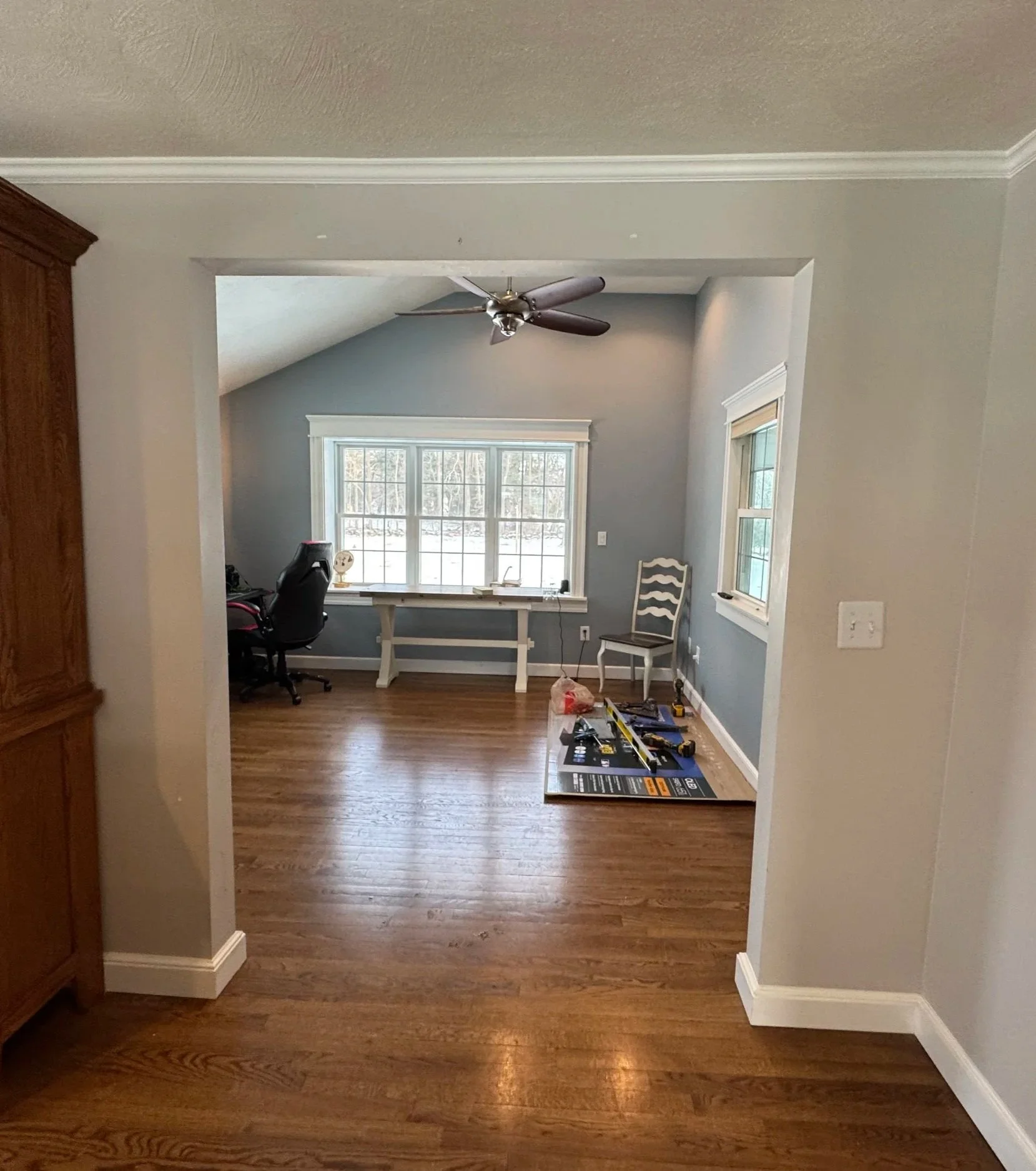 An unfinished room with hardwood floors, light gray walls, and white trim, featuring a ceiling fan, a window with two smaller windows on either side, a desk with a chair, and construction tools on a mat.