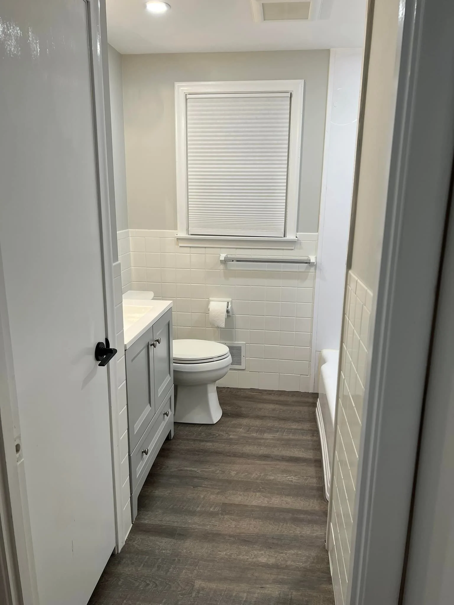A small bathroom with a gray vanity, a white toilet, a window with a closed white blind, and a white bench. The floor is wood-look laminate. The walls are painted light gray with a white tile wainscot.