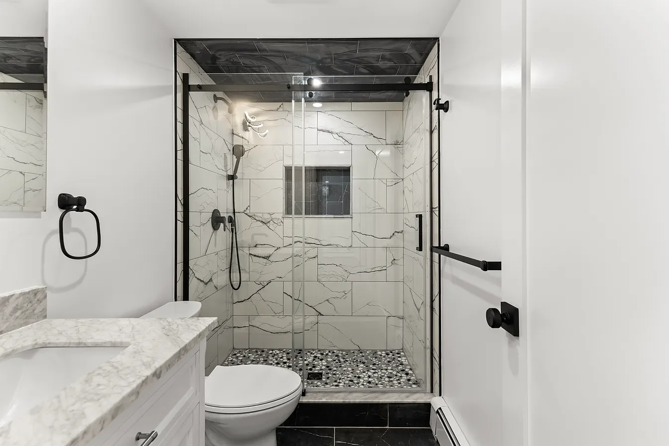 Modern bathroom with a glass shower enclosure featuring black hardware, marble tiles, a pebble shower floor, white walls, a toilet, and a white marble countertop.