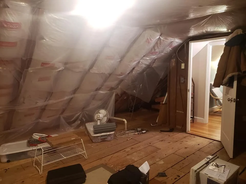 Attic converted into a living space with plastic insulation and various items such as a fan, books, and furniture visible.